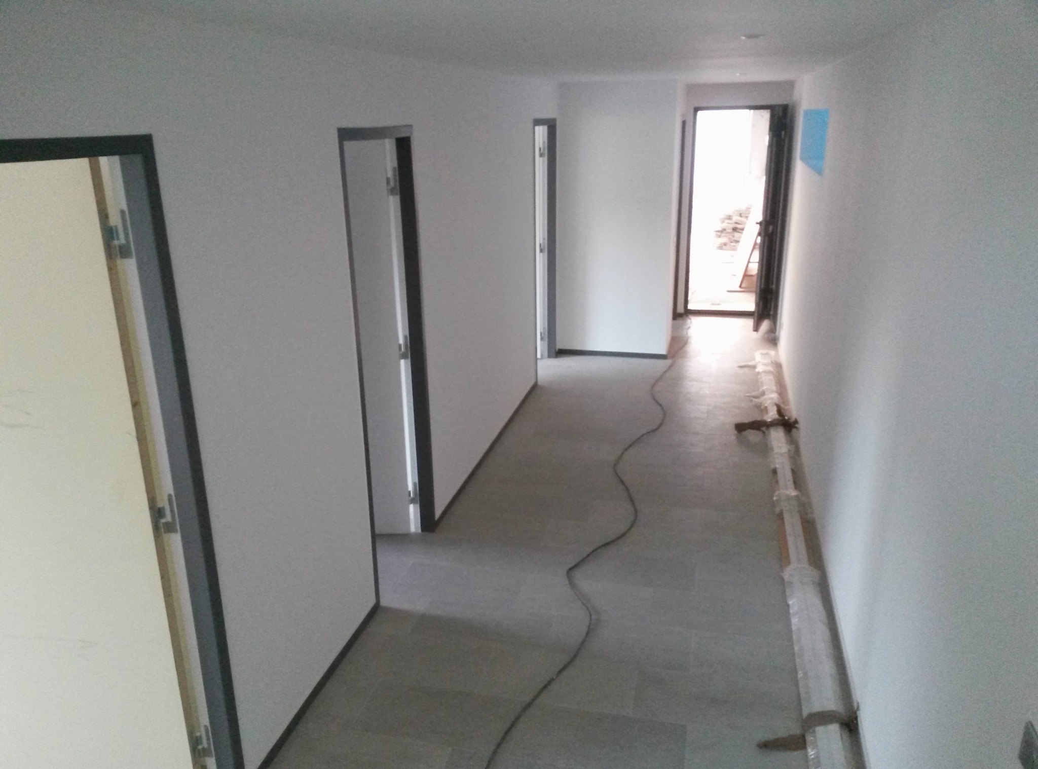 Korytarz po remoncie: szare płytki podłogowe, białe ściany z czarnymi obramowaniami drzwi. Widoczne listwy przypodłogowe w folii i kabel na podłodze.