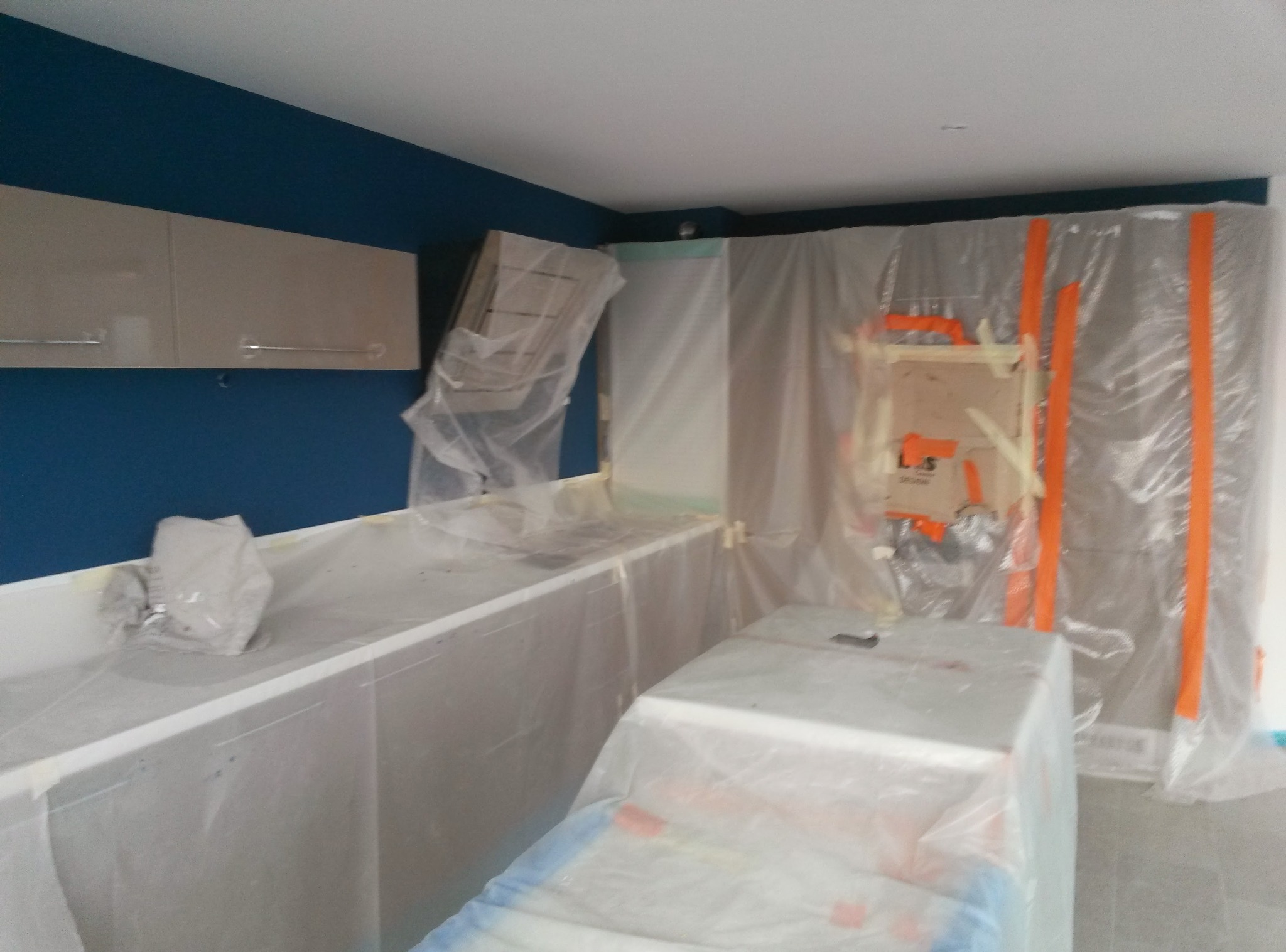 Kuchnia w trakcie zabezpieczania folią malarską przed malowaniem ścian na granatowo. Widoczne szafki i blaty zakryte folią ochronną. Przygotowania do prac malarskich.