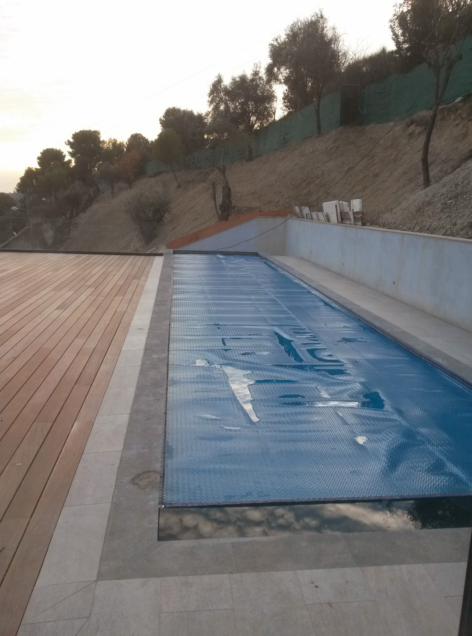 Zadaszony basen zewnętrzny z drewnianym tarasem i kamienną obudową, otoczony wzgórzem z zielenią, w ciepłym świetle zachodzącego słońca.