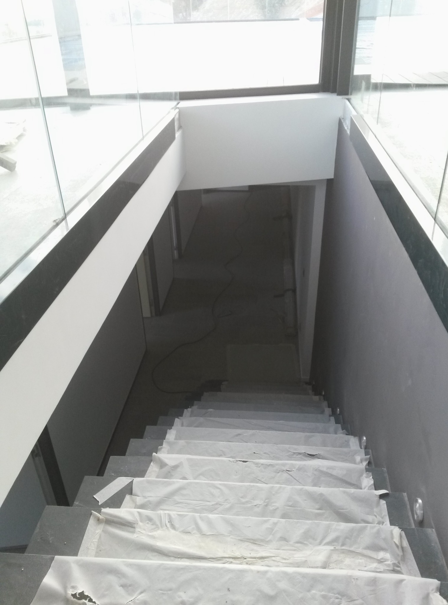 Schody w trakcie zabezpieczania folią ochronną, widok z góry. Szare stopnie, białe ściany, przeszklona balustrada. W tle widoczne pomieszczenie w trakcie prac.