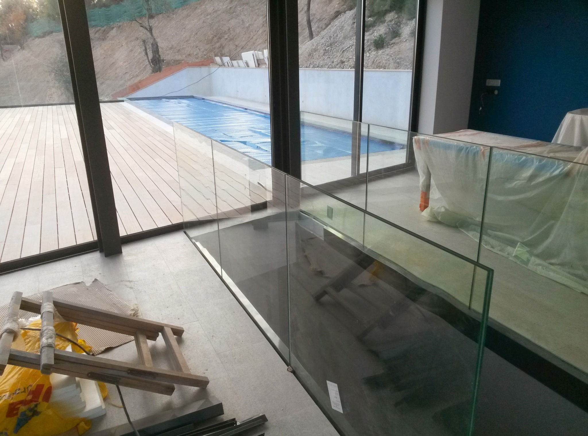 Nowoczesne szklane balustrady wewnętrzne w przestronnym wnętrzu z widokiem na basen. Elementy wykończeniowe w trakcie montażu, widoczne narzędzia i zabezpieczenia.