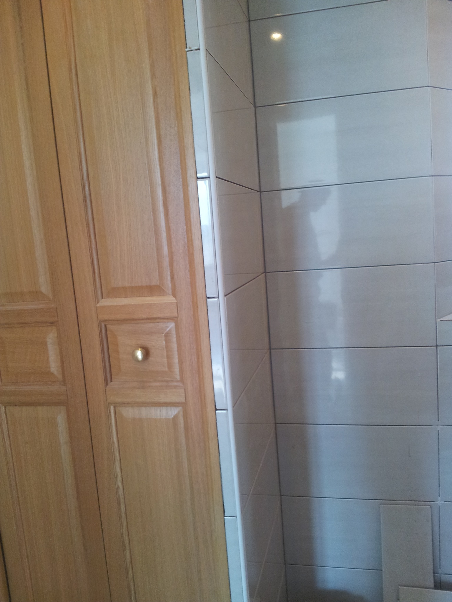 Jasne, kwadratowe płytki w łazience sąsiadują z drewnianymi drzwiami z ozdobnym uchwytem. Widoczny fragment wykończonej ściany.