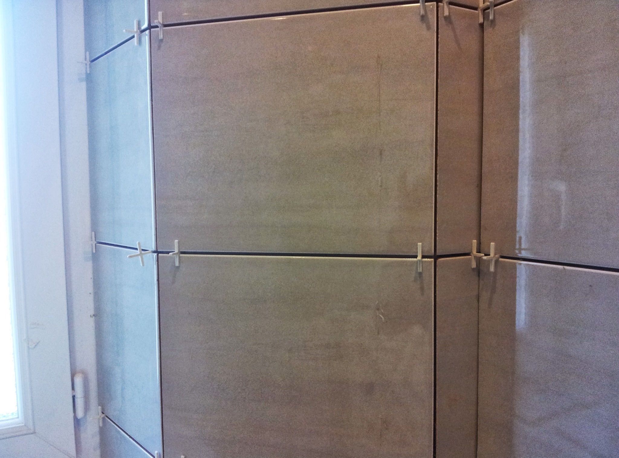 Ściana wyłożona beżowymi płytkami z krzyżykami dystansowymi, widoczne fugi i fragment okna z lewej strony. Ujęcie z bliska, prezentujące detale wykończenia.