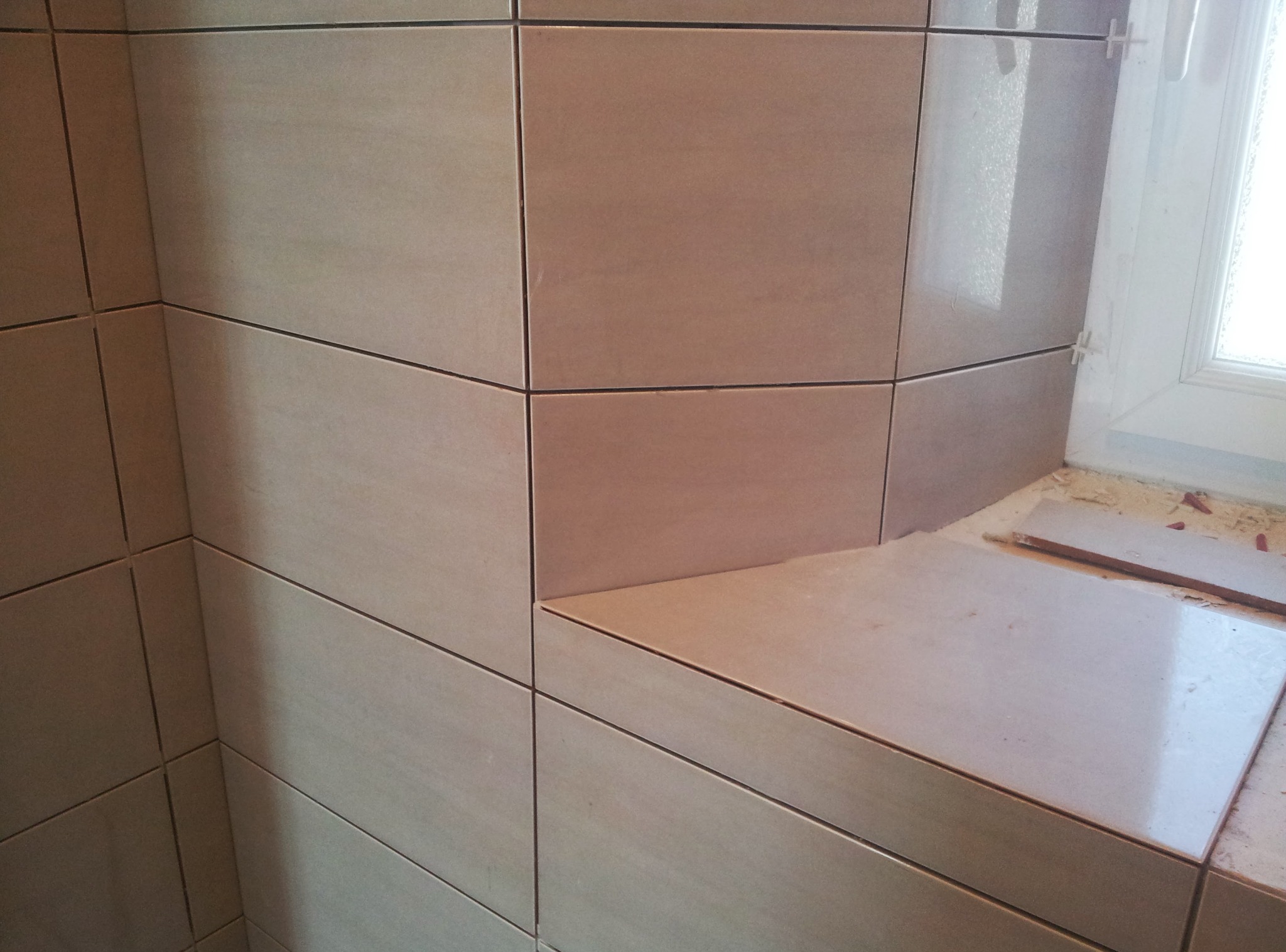 Beżowe, błyszczące płytki ceramiczne ułożone na ścianie i parapecie okiennym w łazience. Widoczne krzyżyki dystansowe. Detal wykończenia.