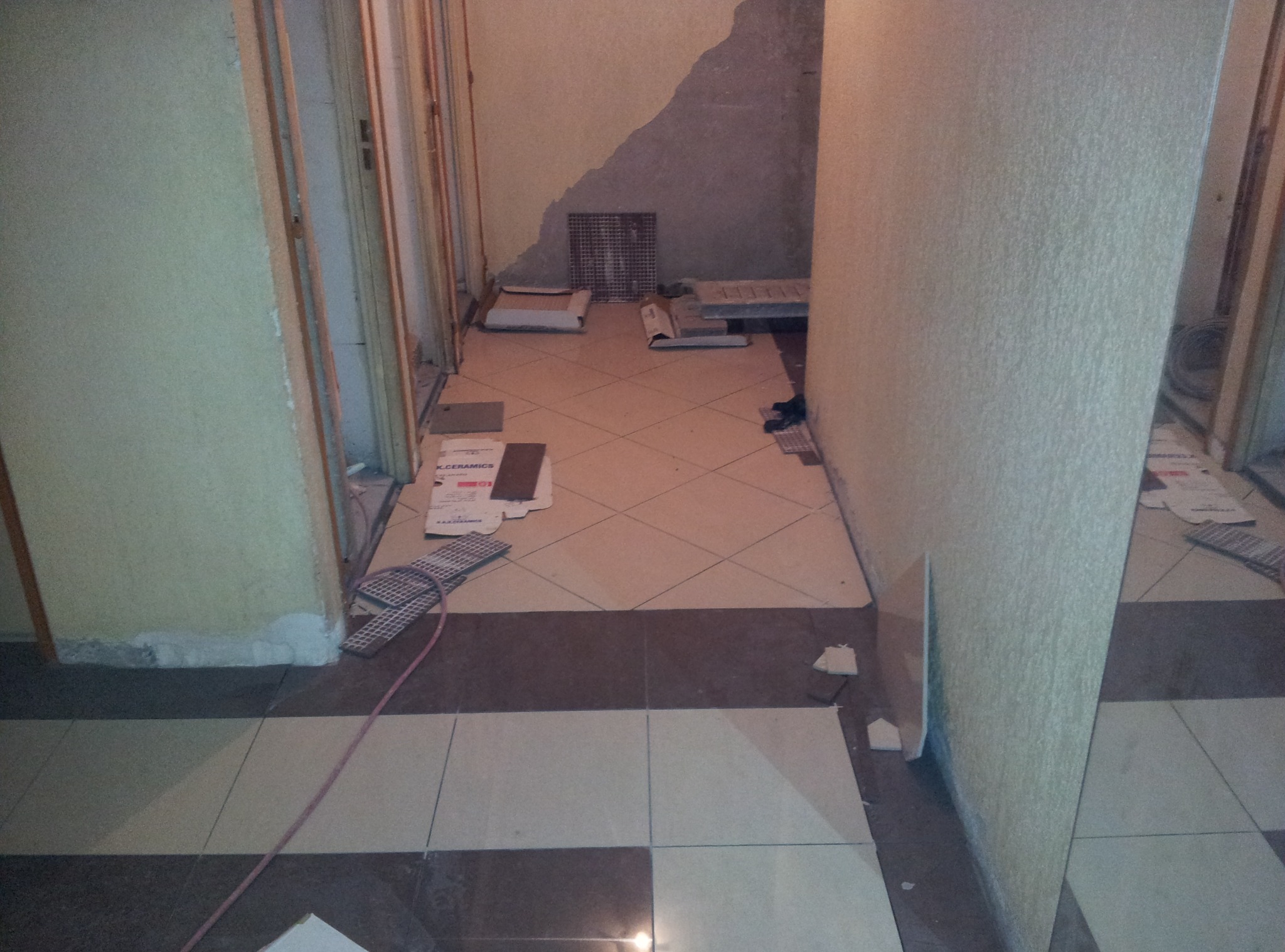 Układanie płytek podłogowych w korytarzu, widoczne materiały i narzędzia glazurnicze, częściowo wykończona podłoga z wzorem w romby i pasy.