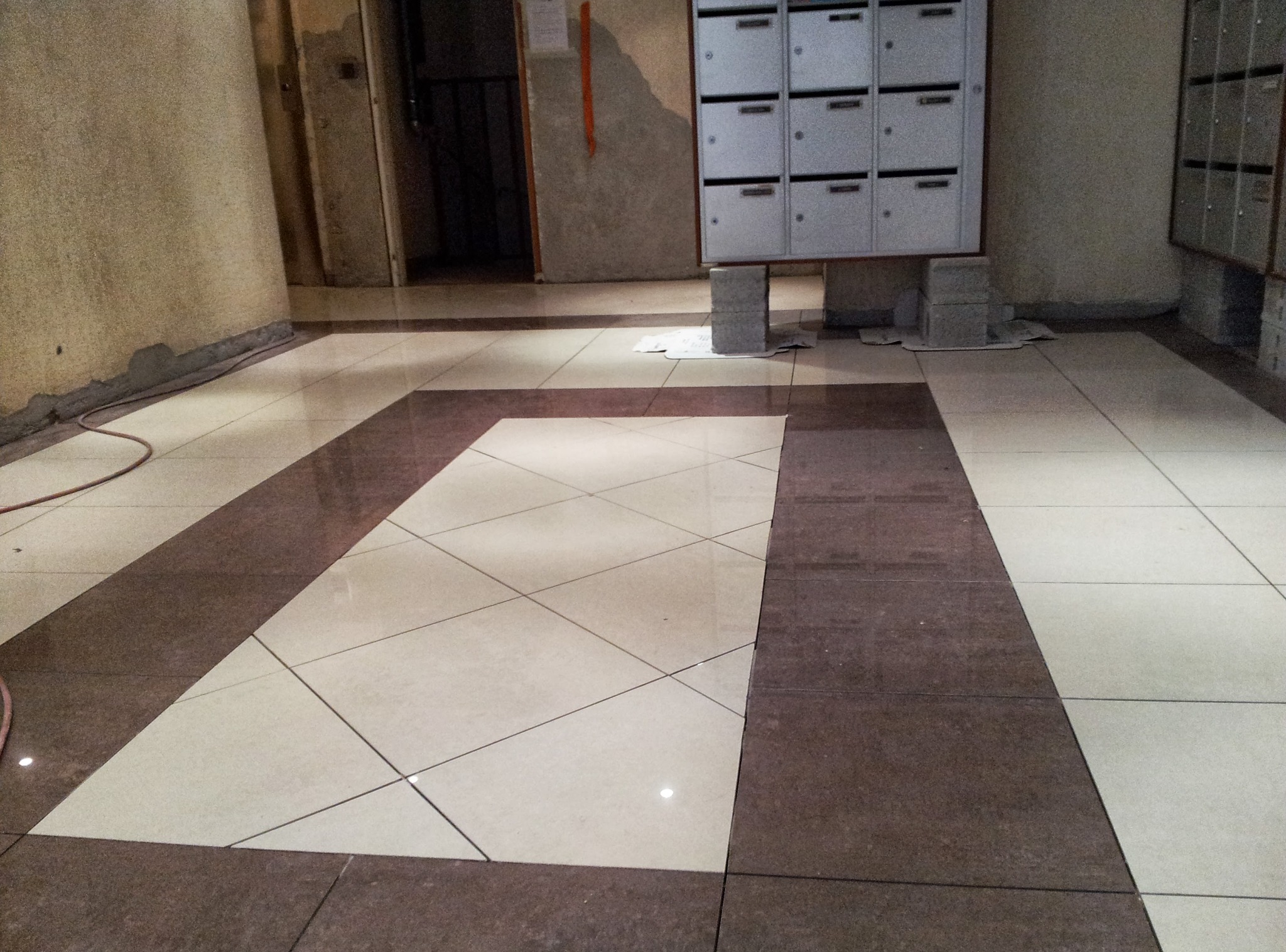 Ułożona podłoga z płytek w korytarzu budynku wielorodzinnego. Brązowe obramowanie z płytek wokół jasnych kwadratów. Skrzynki pocztowe w tle.