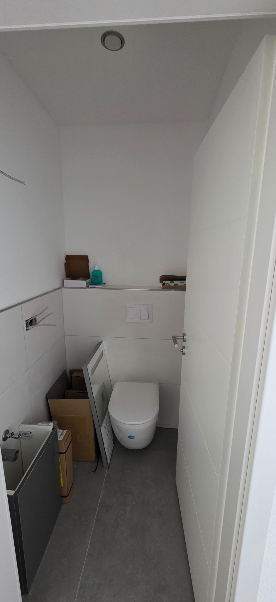 Wnętrze małej, białej łazienki w trakcie wykańczania. Widoczna toaleta, umywalka w zabudowie, białe płytki i otwarte drzwi. Na półce pudełka i akcesoria.