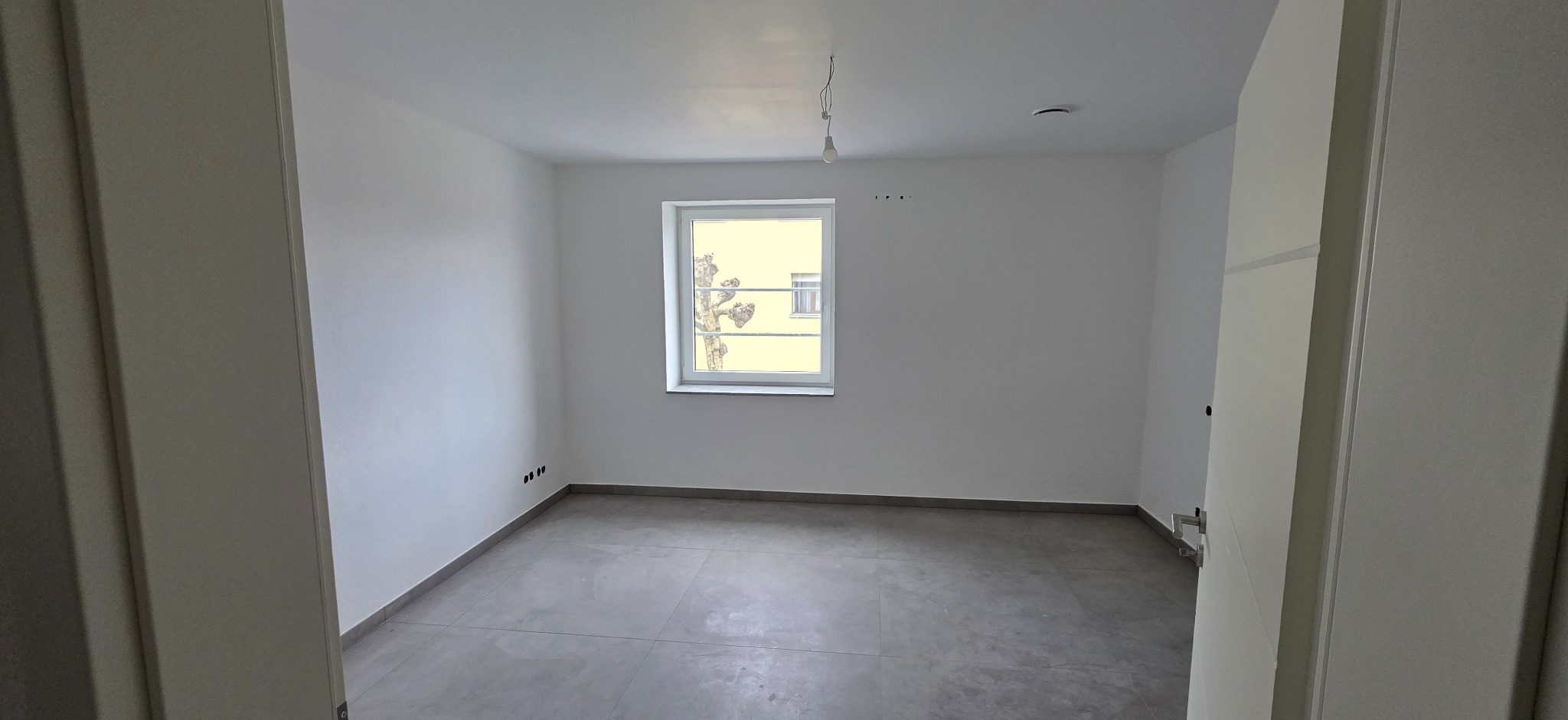 Pusty, odnowiony pokój z szarymi płytkami podłogowymi i białymi ścianami. Widoczne okno z widokiem na drzewo i fragment białych drzwi po prawej stronie kadru.