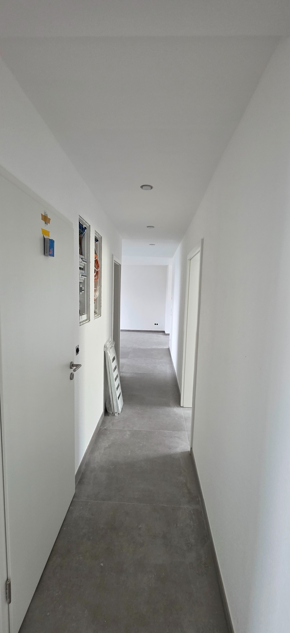 Długi, jasny korytarz z szarą podłogą w nowym budynku. Widoczne białe ściany, drzwi i instalacje elektryczne w trakcie montażu. Minimalistyczny styl wykończenia.