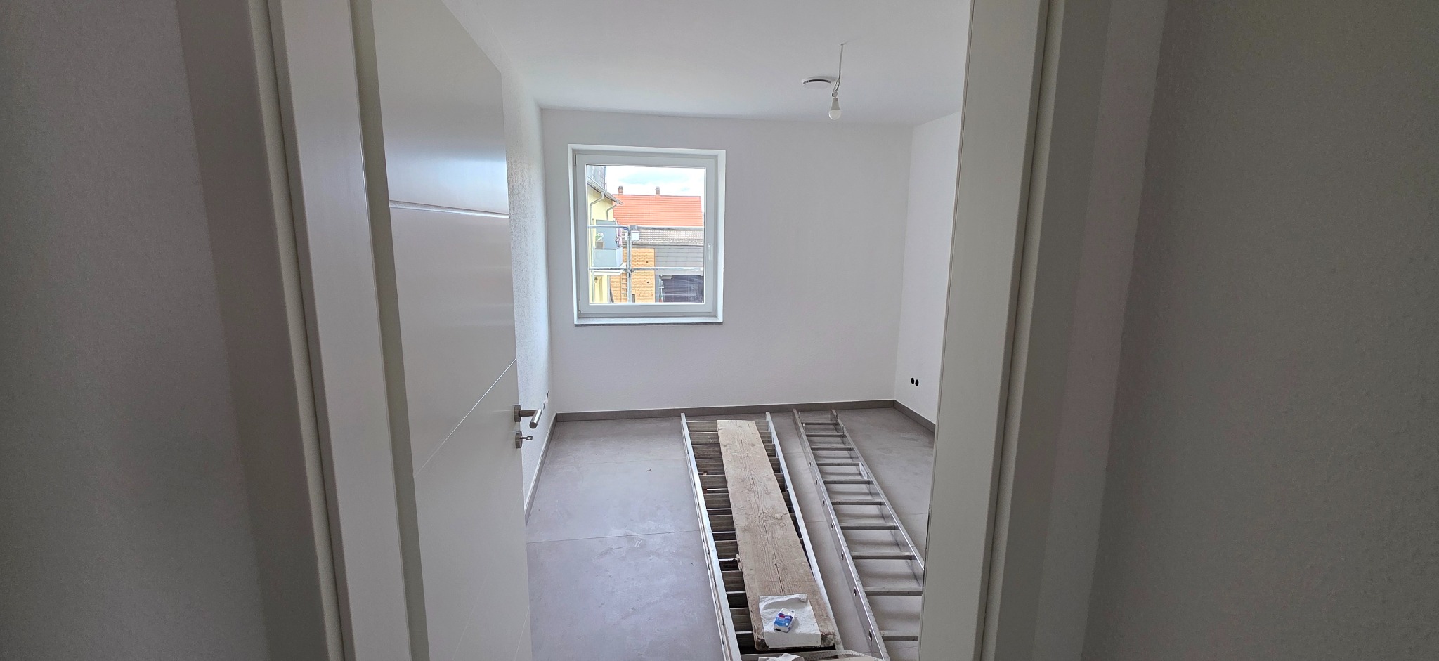 Wnętrze pokoju w trakcie remontu: świeżo pomalowane białe ściany, szara podłoga, drabina i deski na podłodze, widok przez otwarte drzwi.