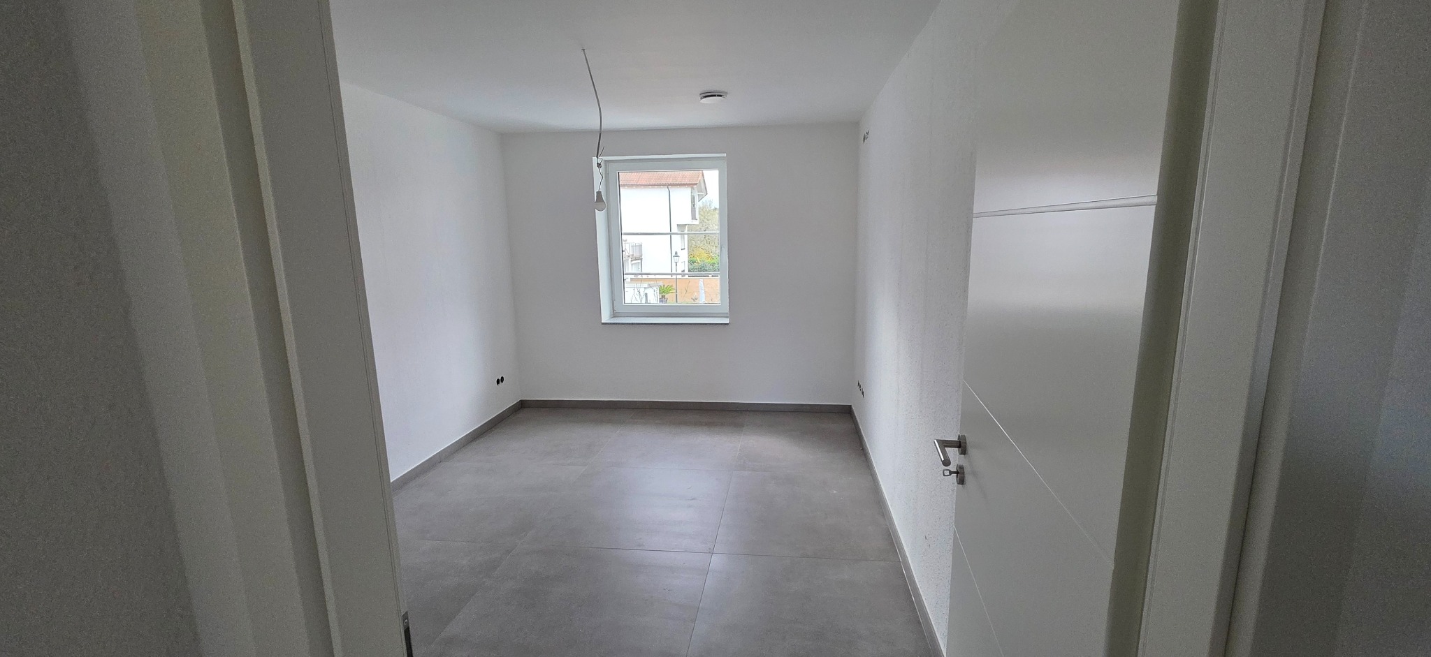 Pusty pokój po remoncie z szarą podłogą z płytek, białymi ścianami i drzwiami. Widoczne okno z widokiem na zewnątrz. Minimalistyczne wykończenie wnętrza.