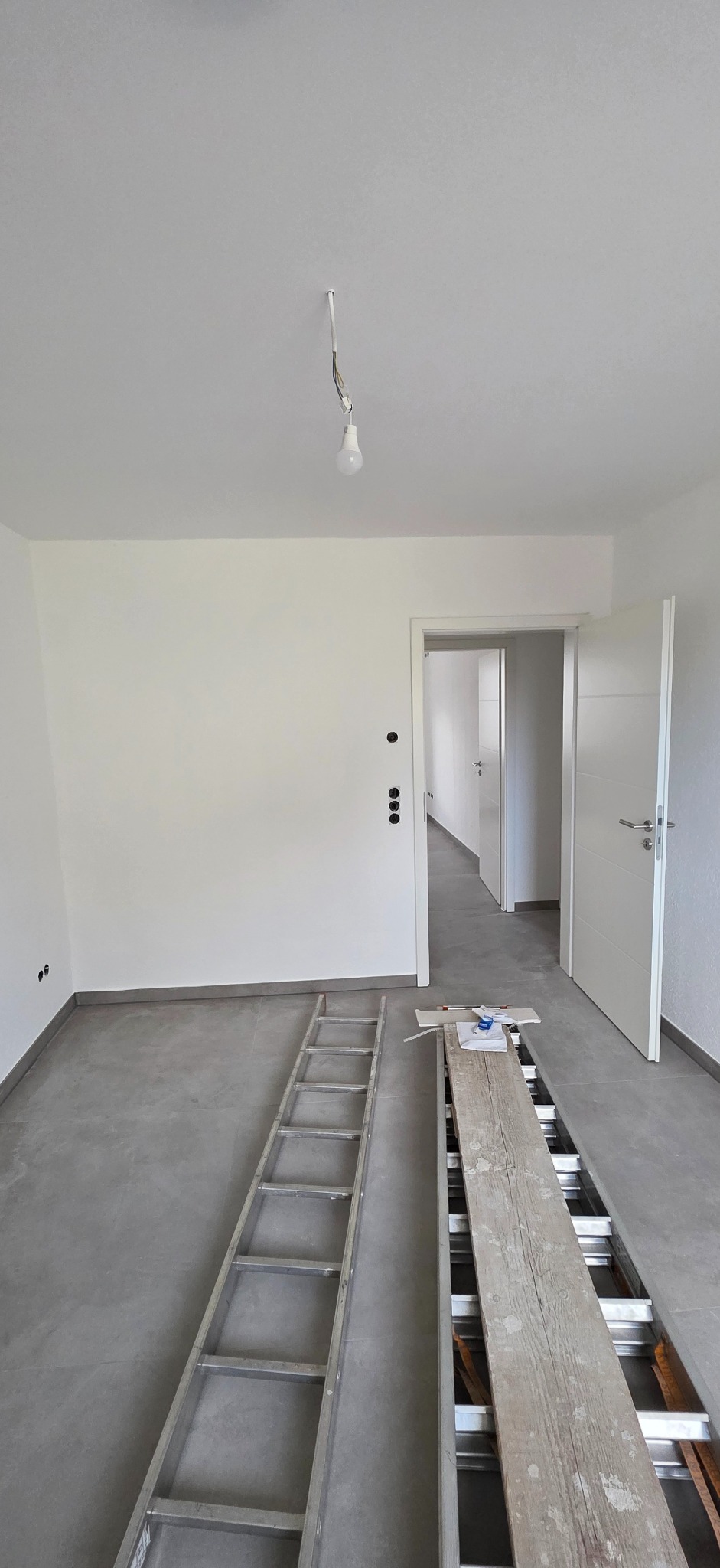Wnętrze po remoncie: białe ściany, szara podłoga, drabina i deska na podłodze. Widoczne gniazdka elektryczne i otwarte drzwi. Żarówka zwisa z sufitu.