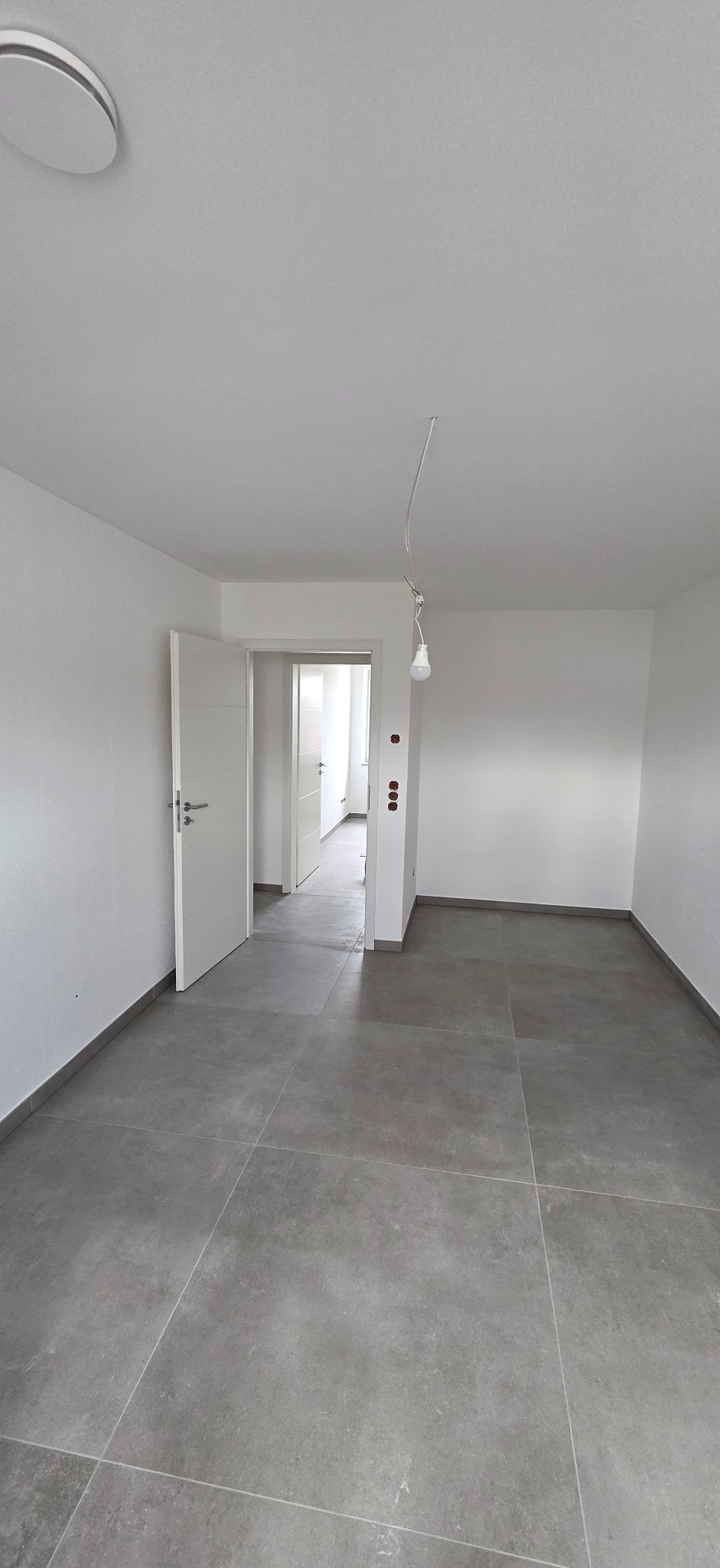 Jasne wnętrze z szarą podłogą z płytek, otwartymi drzwiami i wiszącą żarówką. Minimalistyczny design, świeżo po remoncie, widok na sąsiednie pomieszczenie.