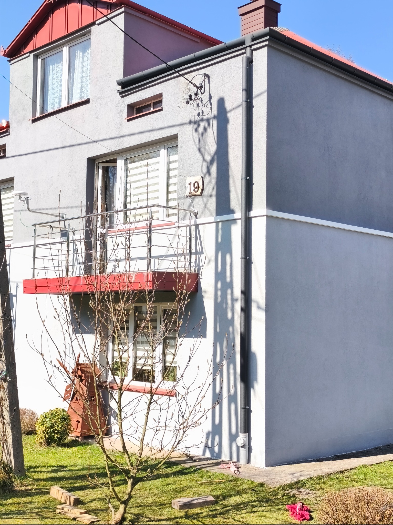 Szary dom z czerwonym dachem i rynnami, balkon z numerem 19, widoczne obróbki blacharskie i fragment ogrodu z drzewem bez liści. Słoneczny dzień.