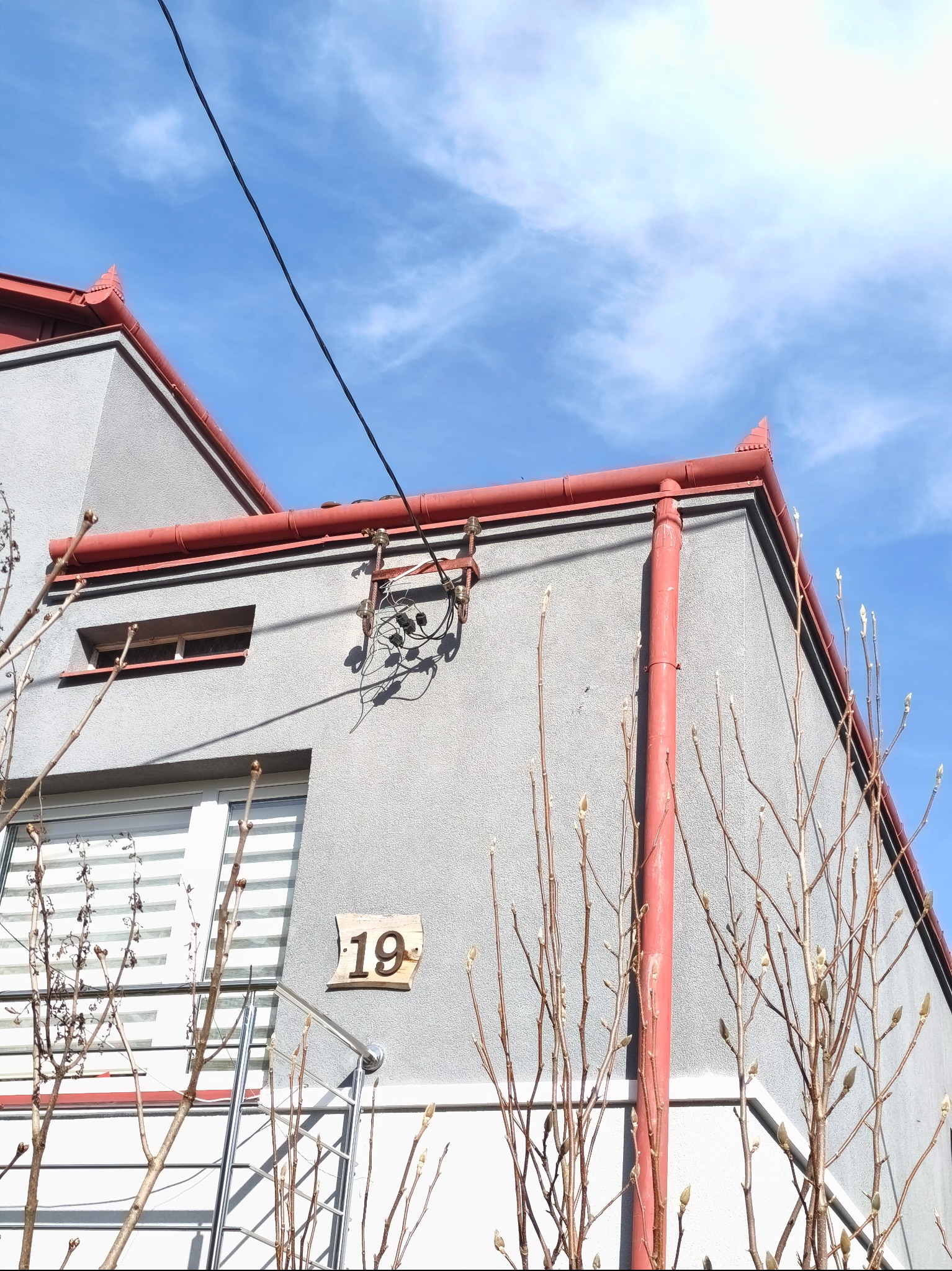 Szary dom z czerwonymi rynnami i numerem 19 na elewacji, widok z dołu na tle błękitnego nieba. Widoczne przewody elektryczne i gałęzie drzew.