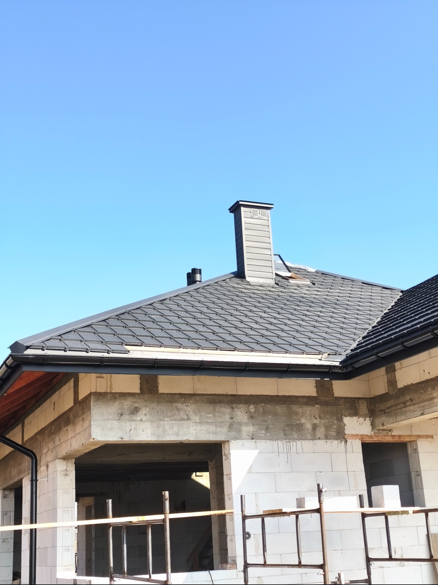 Szary dach z kominem na surowym budynku w trakcie budowy, widoczne rusztowania i niezadaszona konstrukcja. Błękitne niebo w tle.