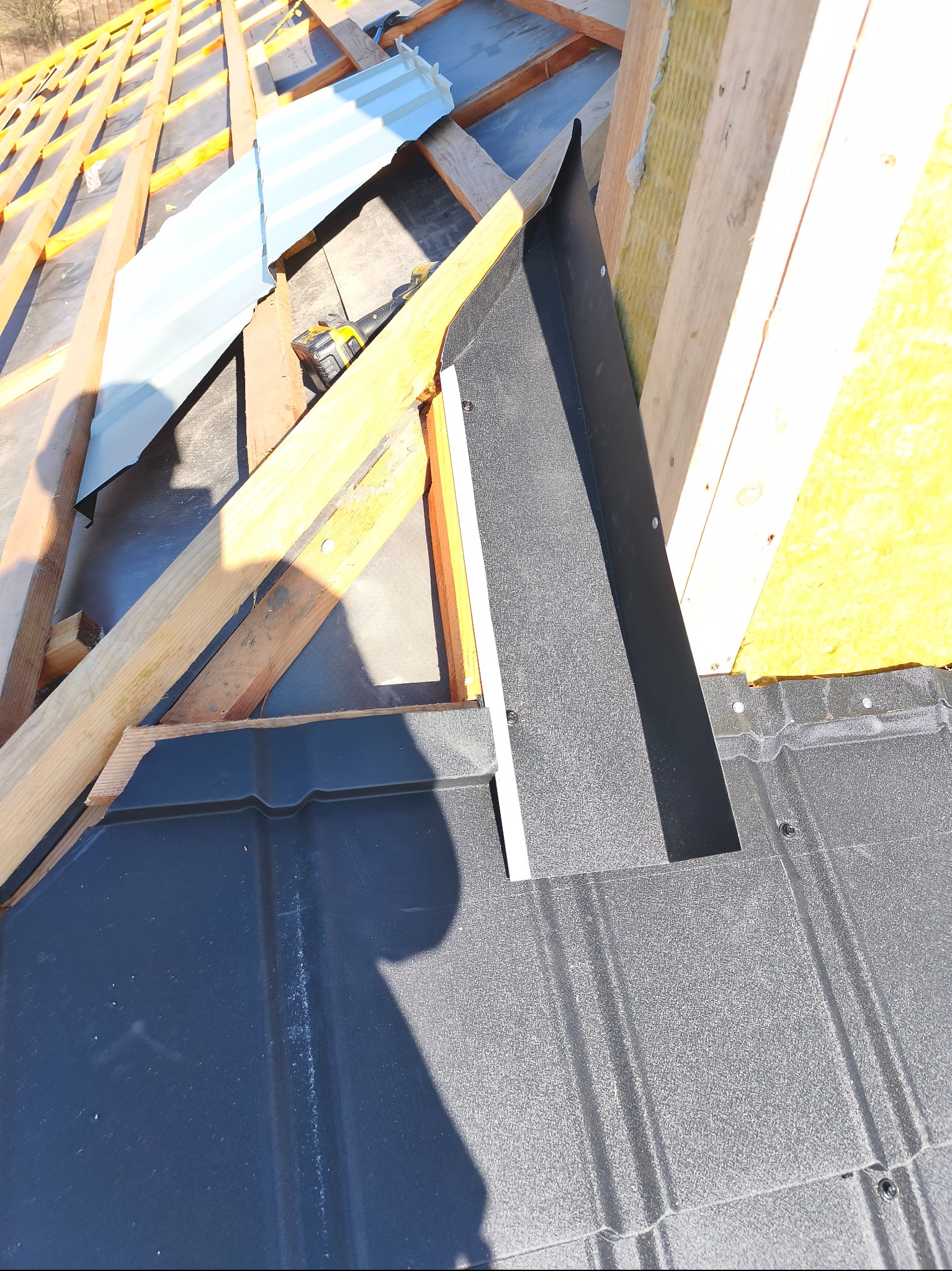 Fragment dachu z czarną blachodachówką, widoczne drewniane elementy konstrukcji, ocieplenie i obróbka blacharska. Ujęcie z góry, w słoneczny dzień.