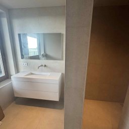 KMK Concept Sp. z o.o. - Nowoczesna łazienka z beżowymi płytkami, białą szafką pod umywalką i dużym lustrem. Widoczny fragment kabiny prysznicowej z brązowymi płytkami.