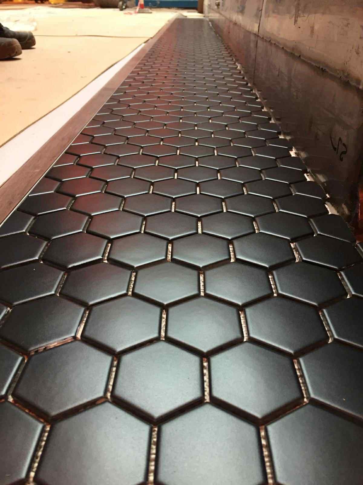 Ułożone, czarne, heksagonalne płytki ceramiczne z widoczną fugą w odcieniu złota, tworzące geometryczny wzór na podłodze. Perspektywa z dołu podkreśla regularność i precyzję ułożenia.