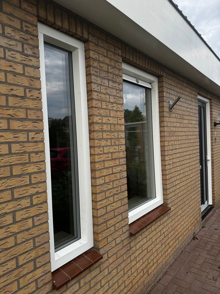 Elewacja budynku z cegły klinkierowej z dwoma oknami PCV z białymi ramami i brązowymi parapetami. Widoczny fragment drzwi wejściowych i rynna.