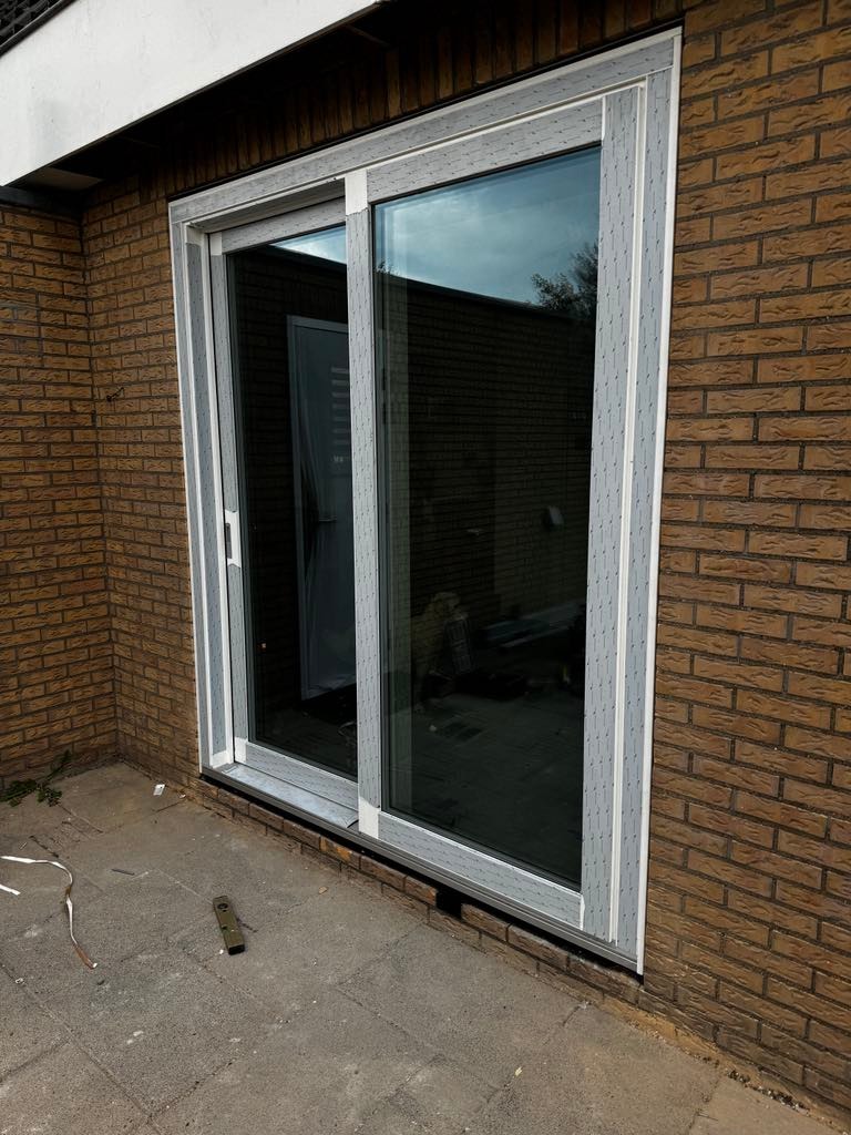 Zamontowane, przesuwne okno tarasowe z szarymi ramami w murze z cegły. Widoczny poziomica na posadzce z kostki brukowej.