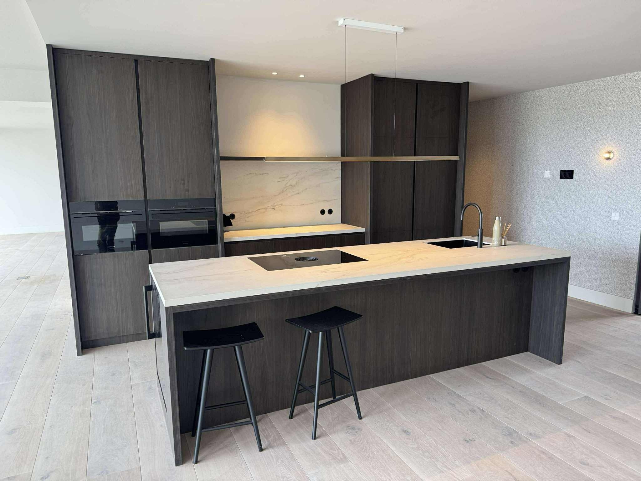 Nowoczesna kuchnia z ciemnymi drewnianymi szafkami, marmurowym blatem i dwoma czarnymi stołkami barowymi na jasnej drewnianej podłodze. Minimalistyczny design.