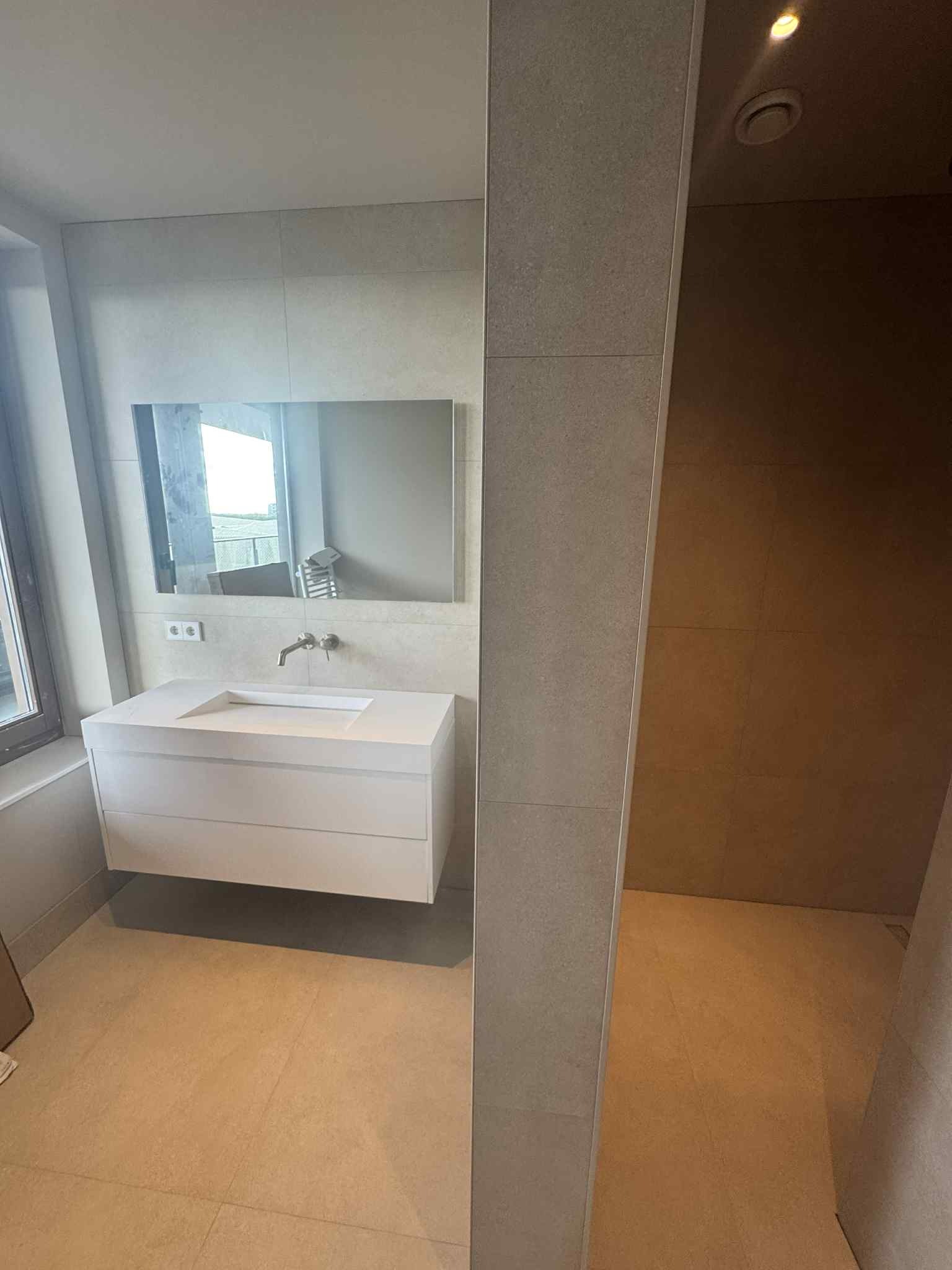 Nowoczesna łazienka z beżowymi płytkami, białą szafką pod umywalką i dużym lustrem. Widoczny fragment kabiny prysznicowej z brązowymi płytkami.