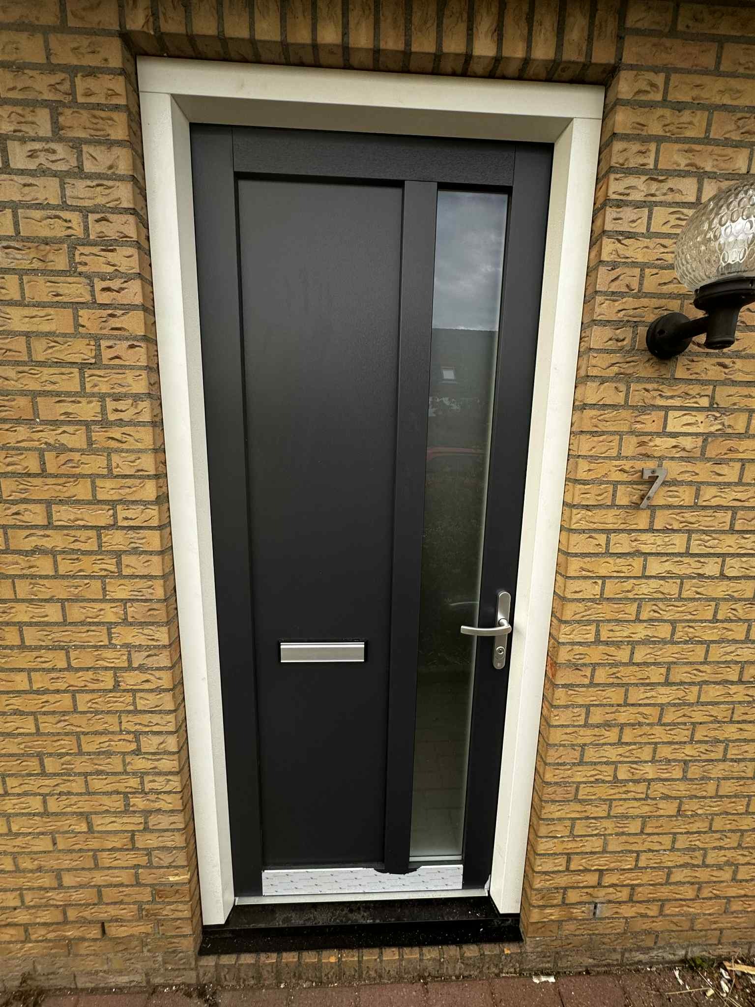 Ciemne drzwi wejściowe z szybą, osadzone w białej futrynie, na tle ściany z żółtej cegły. Widoczny numer domu '7' i zewnętrzna lampa.
