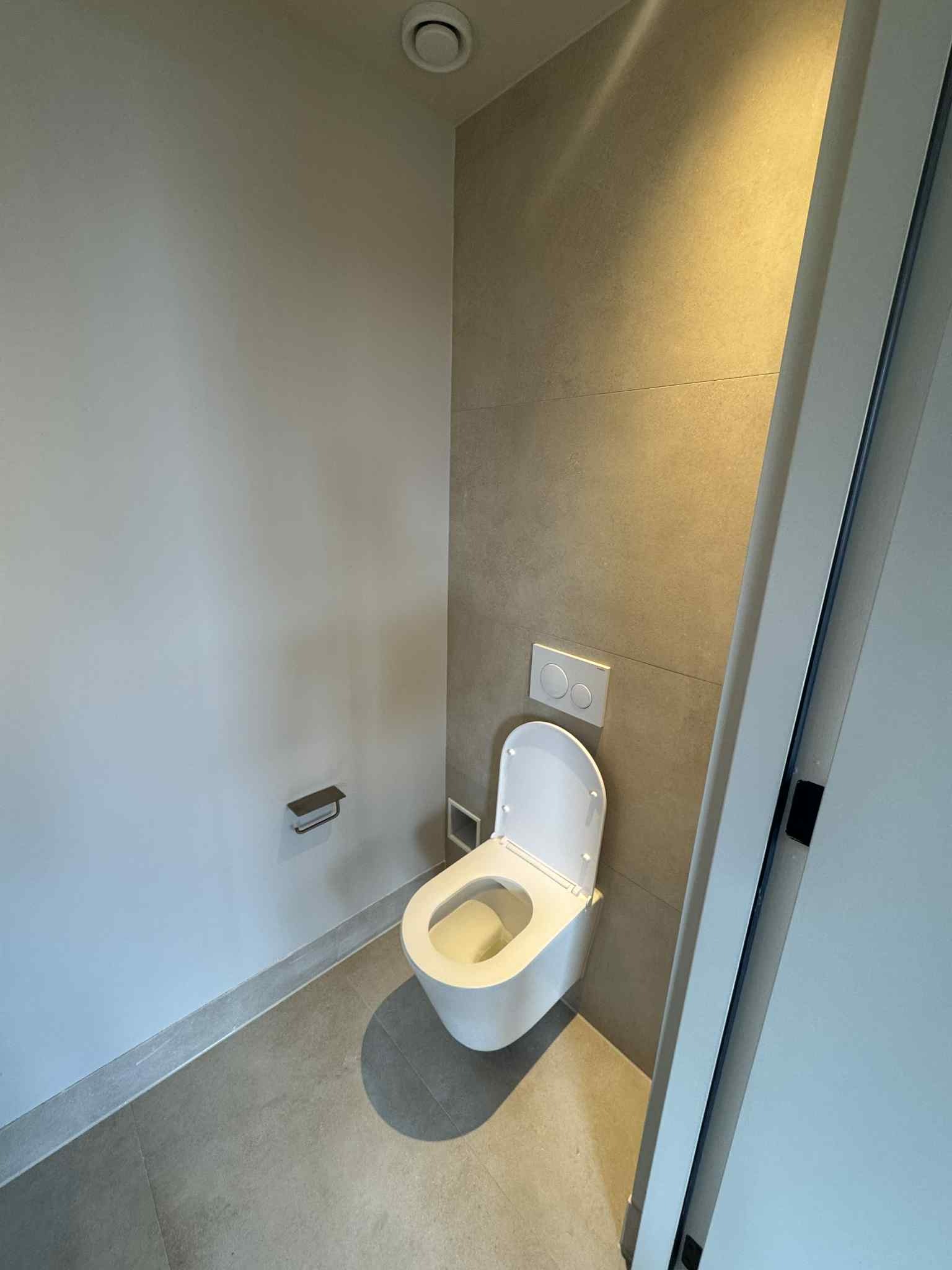 Nowoczesna toaleta podwieszana w minimalistycznej łazience z szarą płytką ścienną i podłogową. Białe ściany, widoczny fragment drzwi. Oświetlenie sufitowe.