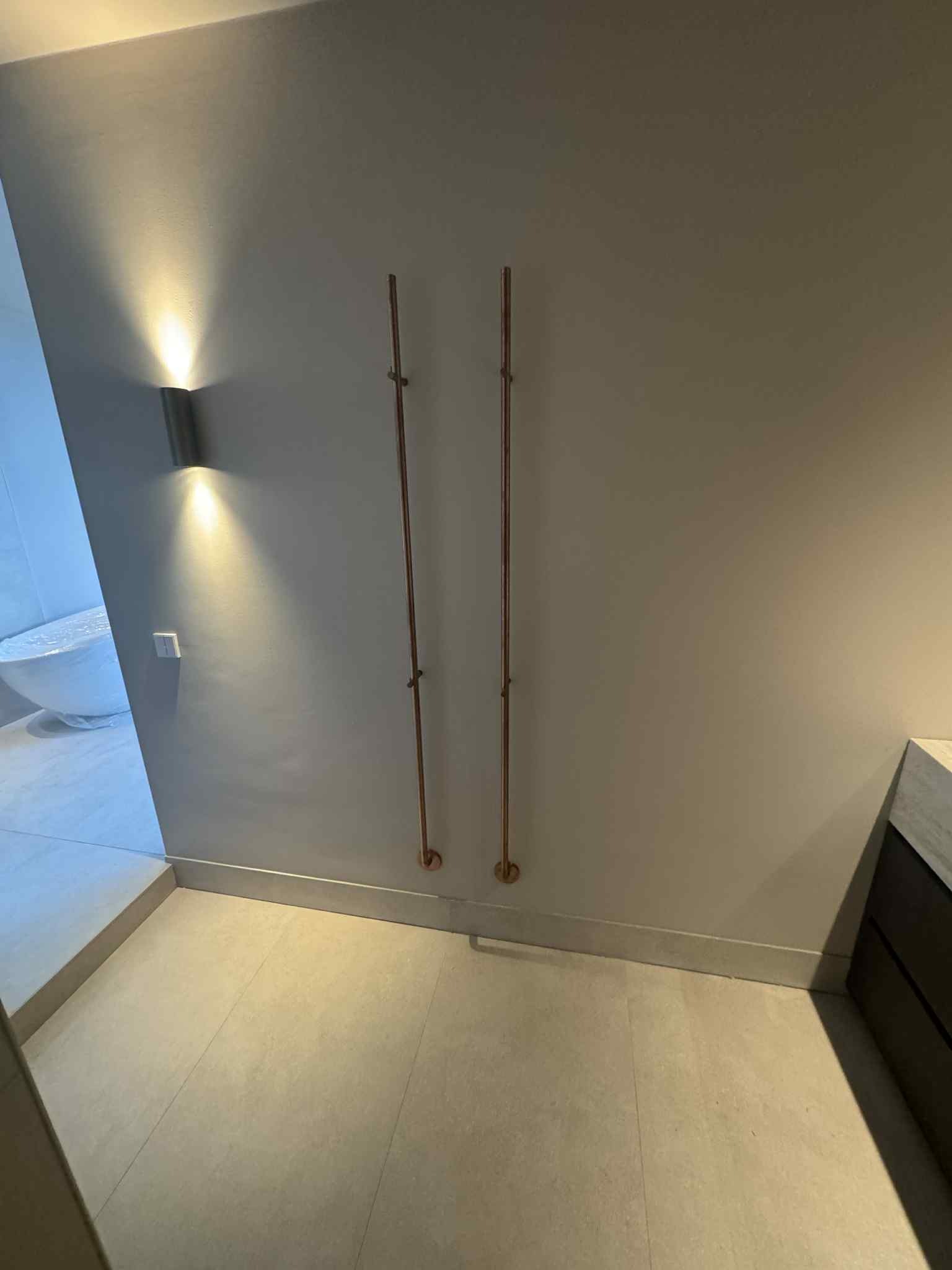 Minimalistyczna łazienka z miedzianymi rurami grzewczymi na ścianie, oświetlona nowoczesnym kinkietem. Jasne płytki podłogowe i fragment wanny w tle.