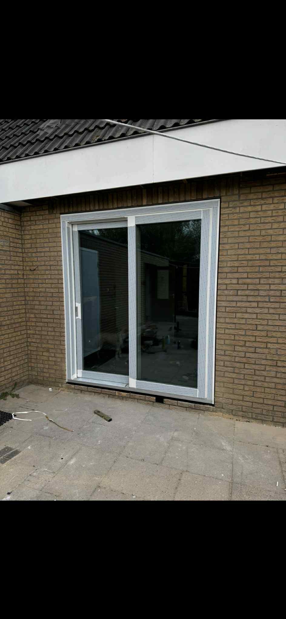 Nowe, przesuwne okno tarasowe z jasną ramą w elewacji z cegły klinkierowej. Widoczne detale wykończenia i fragment tarasu z szarych płyt.