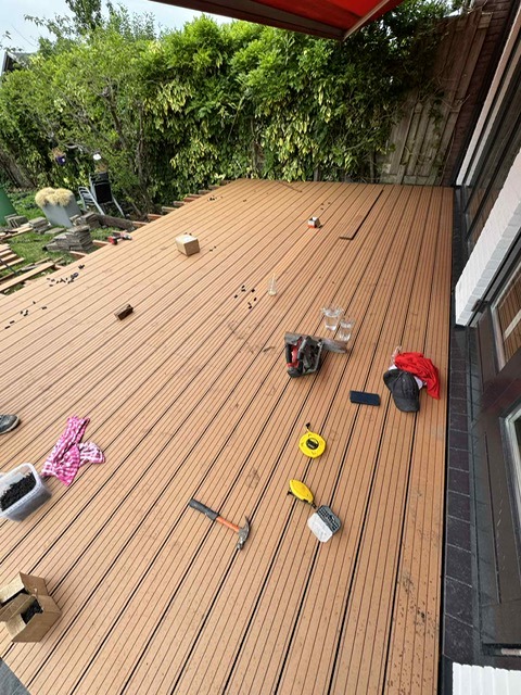 Montaż deski kompozytowej na tarasie w Kielcach. Widoczne narzędzia: piła, młotek, miarka. Częściowo ukończona podłoga tarasu z desek w odcieniach brązu.