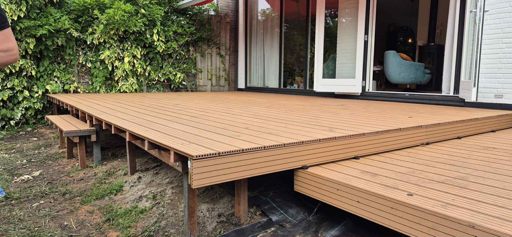 Nowoczesny taras z kompozytowych desek w odcieniach brązu, z niskim stopniem i siedziskiem, przylegający do domu z widokiem na wnętrze przez otwarte drzwi.