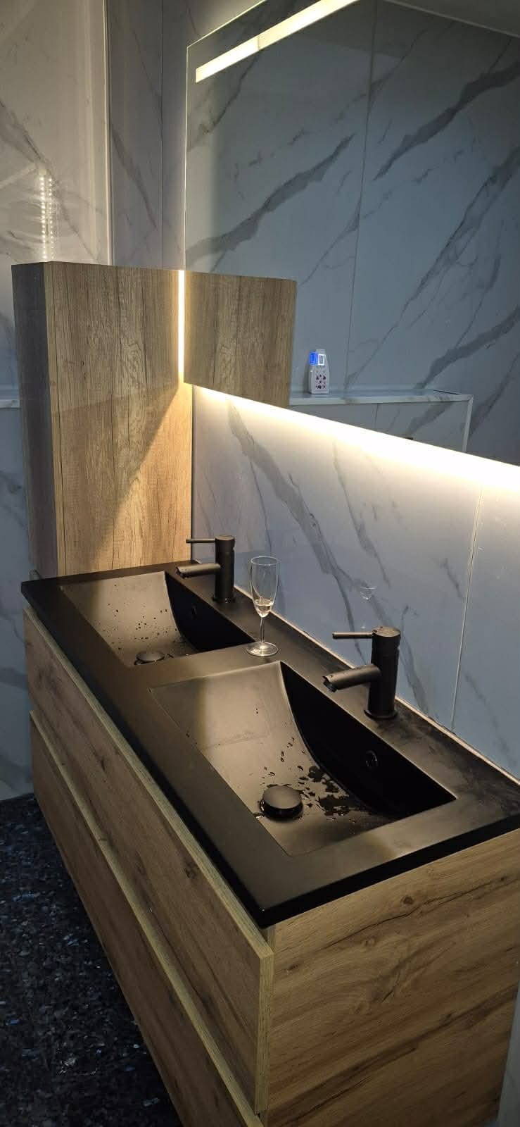 Nowoczesna łazienka z podwójną, czarną umywalką na szafce z drewnopodobnym frontem. Marmurowe płytki na ścianach i subtelne oświetlenie LED.