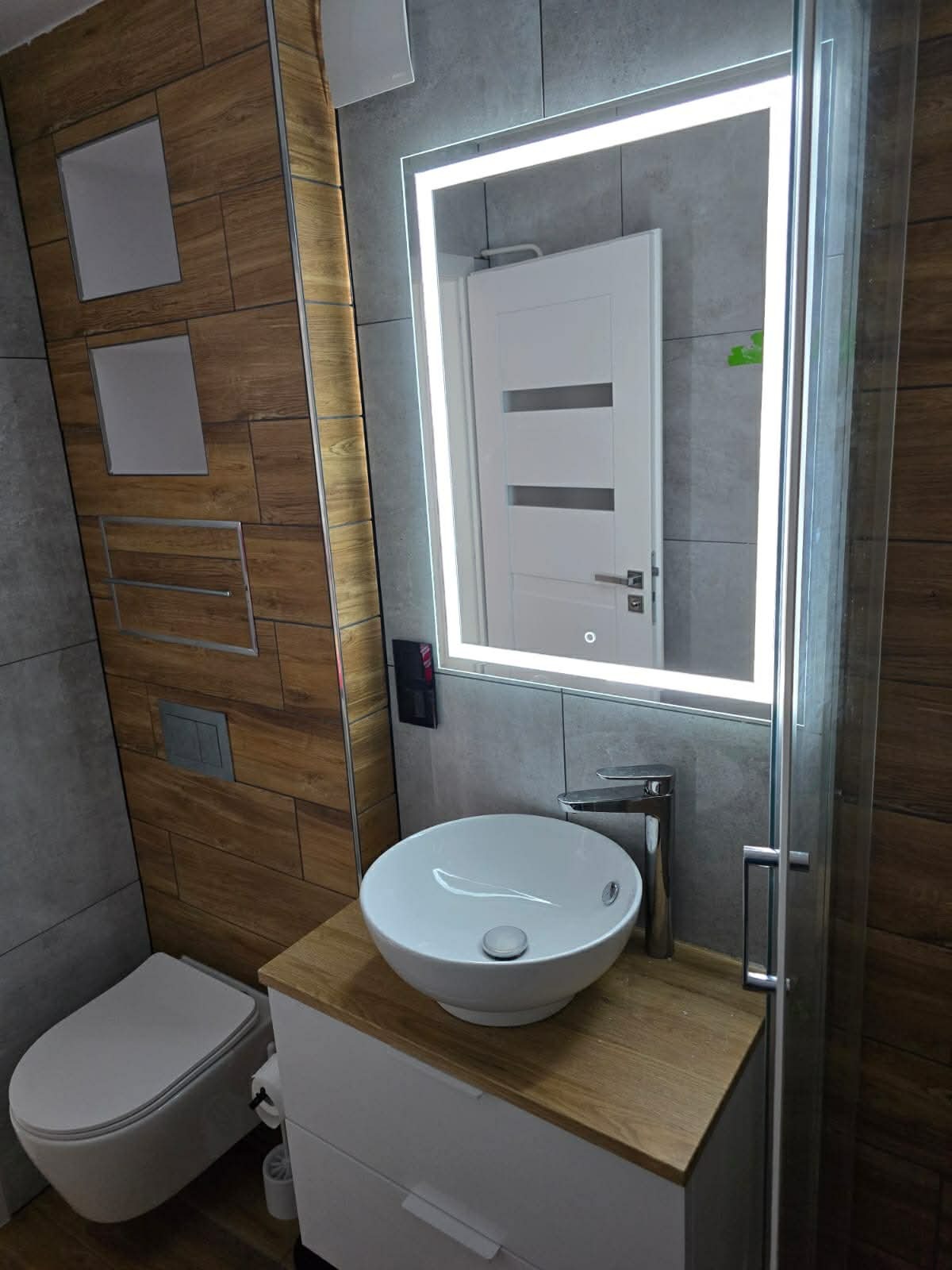 Nowoczesna łazienka z umywalką nablatową, podświetlanym lustrem i drewnopodobnymi płytkami. Widoczny fragment kabiny prysznicowej i wisząca toaleta.