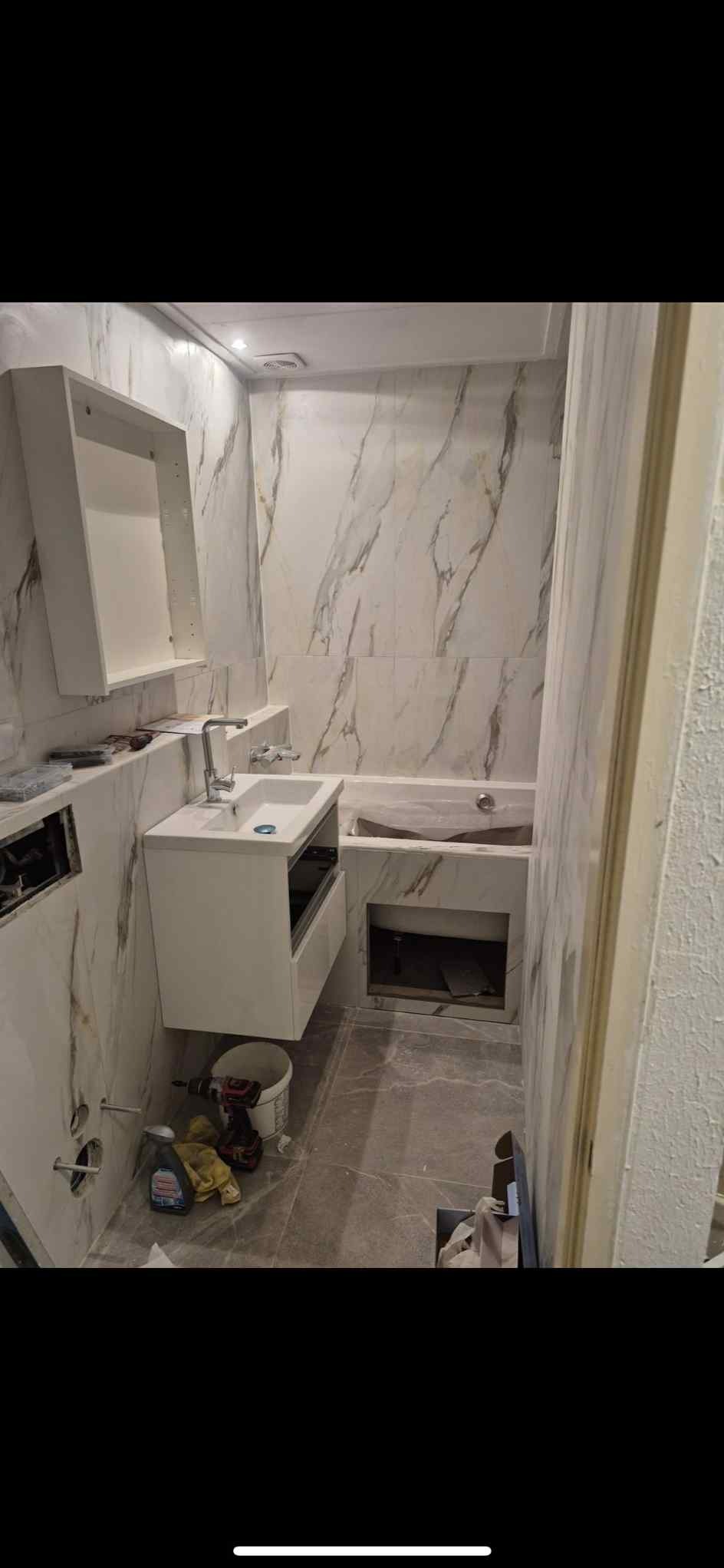 Remont łazienki w trakcie: białe marmurowe płytki, biała szafka pod umywalką, wanna w zabudowie. Widoczne narzędzia i materiały budowlane.