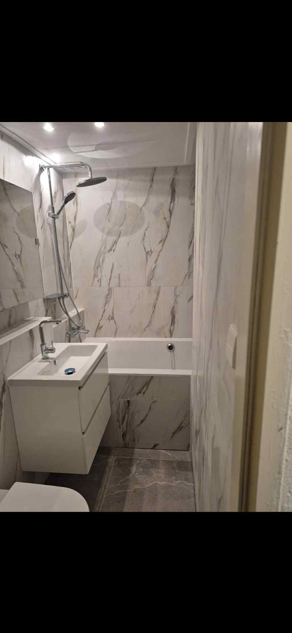 Wykończona łazienka z wanną, umywalką z szafką i marmurowymi płytkami na ścianach. Nowoczesny design, szara podłoga, oświetlenie punktowe.