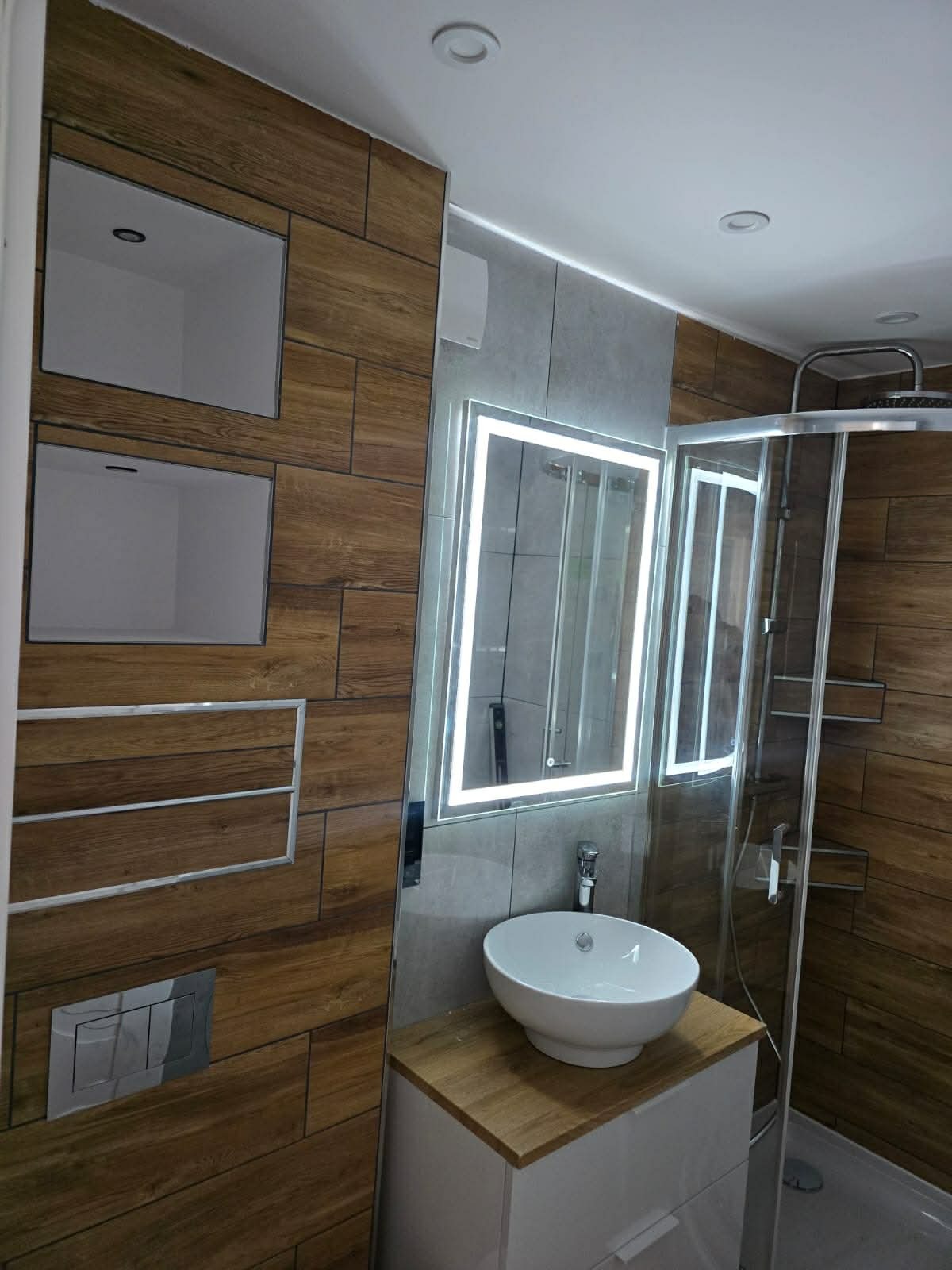 Nowoczesna łazienka z drewnopodobnymi płytkami, podświetlanym lustrem i kabiną prysznicową typu walk-in. Umywalka nablatowa na szafce z blatem imitującym drewno.