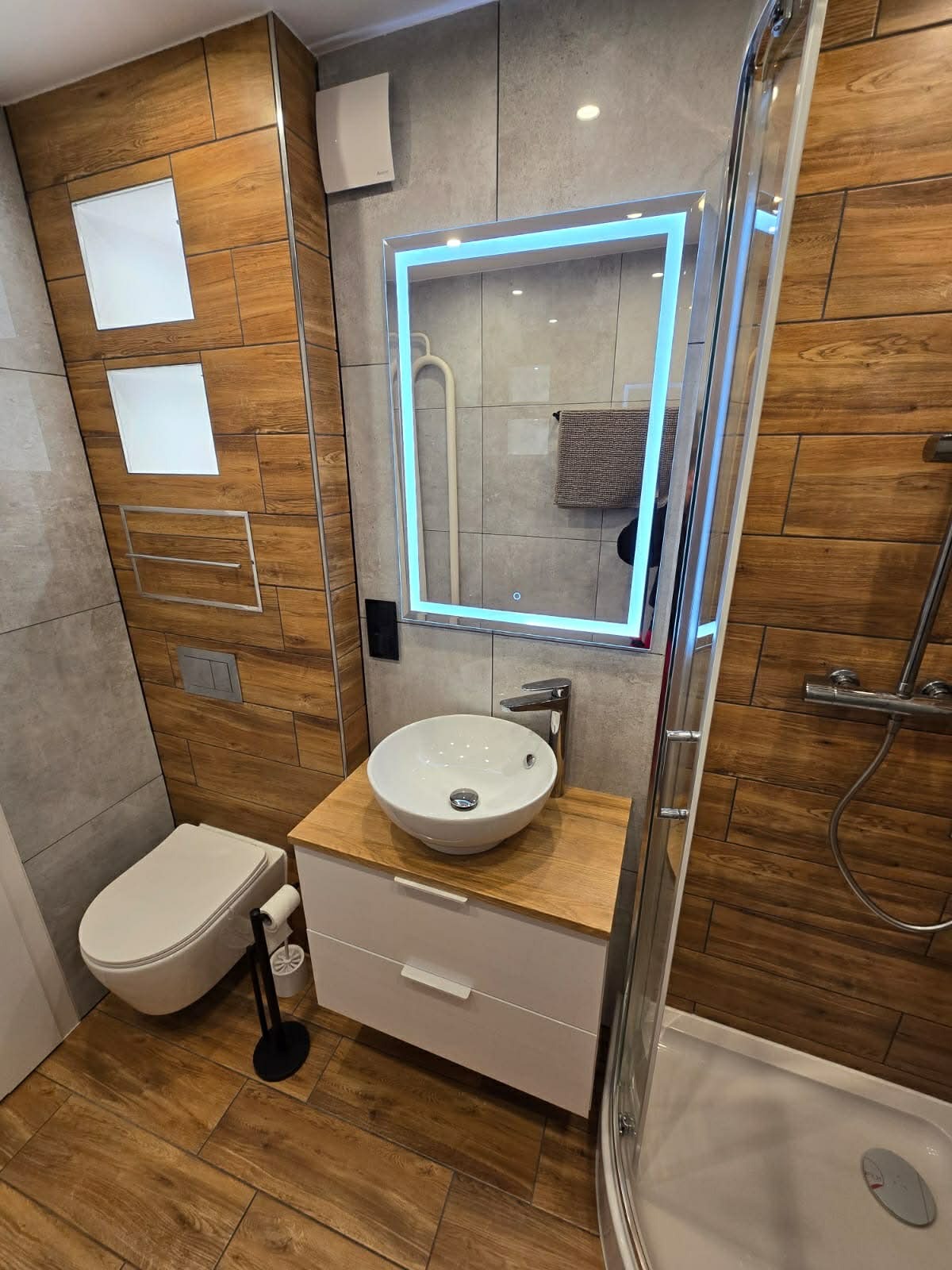 Nowoczesna łazienka po remoncie: drewnopodobne płytki, podświetlane lustro, biała umywalka nablatowa i kabina prysznicowa z transparentnego szkła.