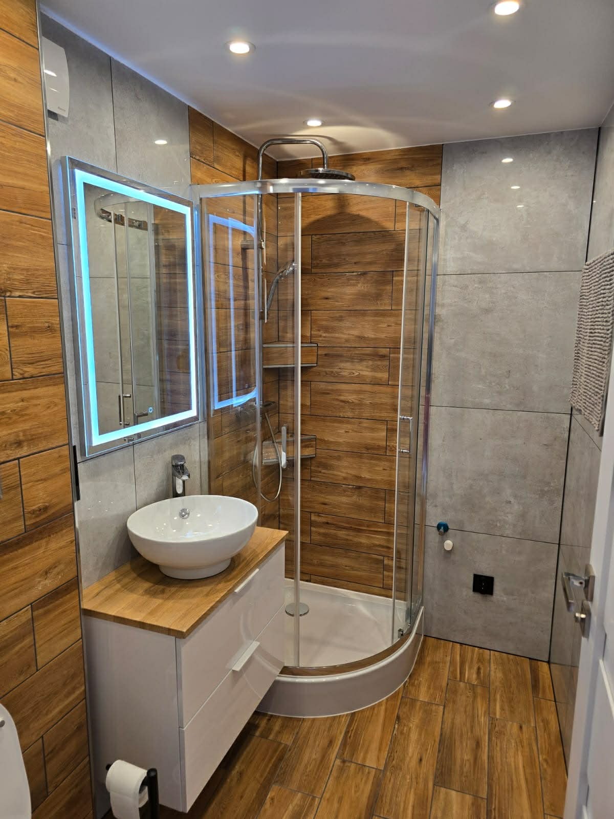 Nowoczesna łazienka po remoncie: kabina prysznicowa z chromowanymi profilami, umywalka nablatowa na szafce i podświetlane lustro. Płytki imitujące drewno i szarość.