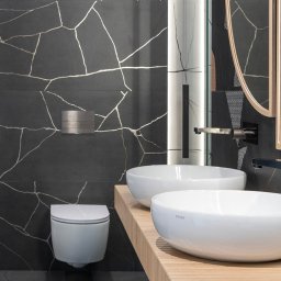 NOWATOM Tomasz Międła - Nowoczesna łazienka z czarnymi płytkami imitującymi marmur ze złotymi żyłami, dwie białe umywalki na drewnianym blacie i podwieszana toaleta.