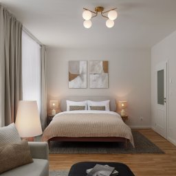 NOWATOM Tomasz Międła - Jasna sypialnia w neutralnych barwach: drewniana podłoga, beżowe zasłony, łóżko z narzutą, sofa i lampy. Minimalistyczne obrazy nad łóżkiem, lustro w czarnej ramie.