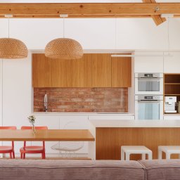 Tomasz Międła - Jasna kuchnia z drewnianymi akcentami: szafki, blat, stół i lampy. Ceglana ściana nad blatem. Minimalistyczny design, nowoczesne oświetlenie, widoczne belki stropowe.
