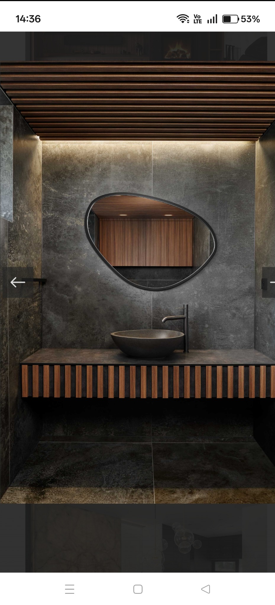 Nowoczesna łazienka z umywalką w kształcie misy, lustrem o nieregularnym kształcie i drewnianymi akcentami na tle szarych płytek. Oświetlenie LED podkreśla detale.