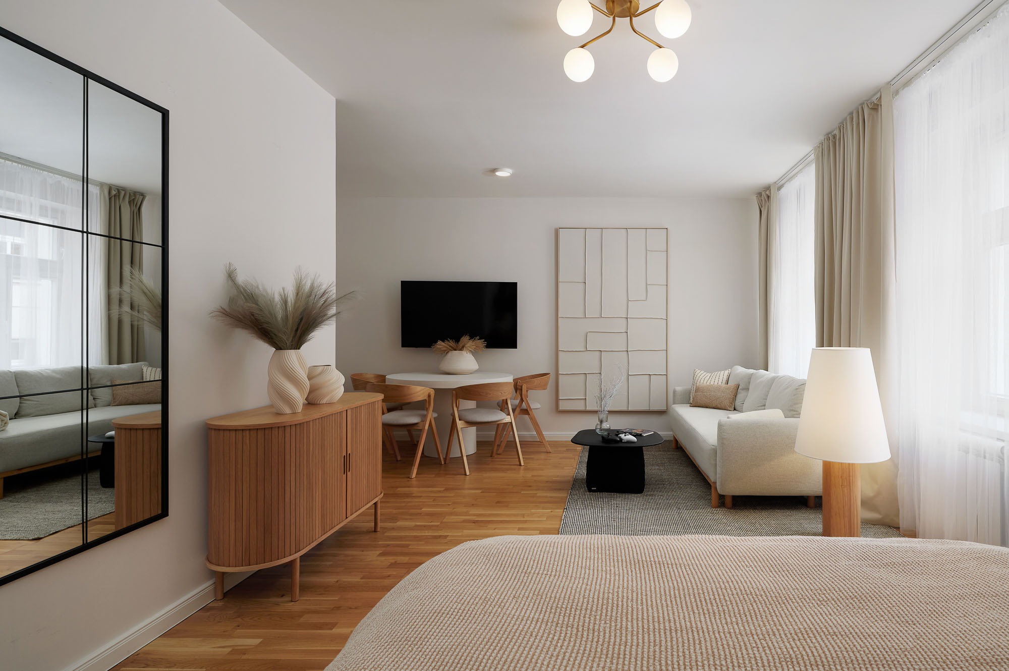 Jasne, przestronne wnętrze mieszkania w stylu skandynawskim. Widoczny salon z sofą, stolikiem kawowym, jadalnia z okrągłym stołem i drewnianymi krzesłami oraz komoda z dekoracjami.