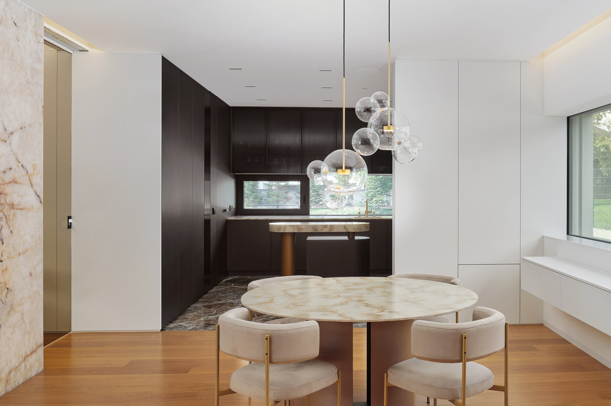Eleganckie wnętrze: okrągły stół z krzesłami, nowoczesna lampa wisząca z kul, zabudowa kuchenna w ciemnym drewnie, marmurowe wykończenia i drewniana podłoga.