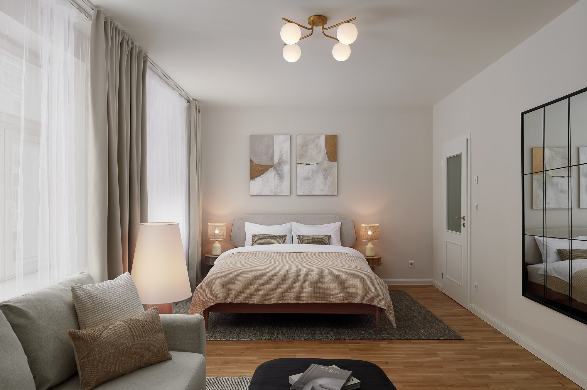 Jasna sypialnia w neutralnych barwach: drewniana podłoga, beżowe zasłony, łóżko z narzutą, sofa i lampy. Minimalistyczne obrazy nad łóżkiem, lustro w czarnej ramie.