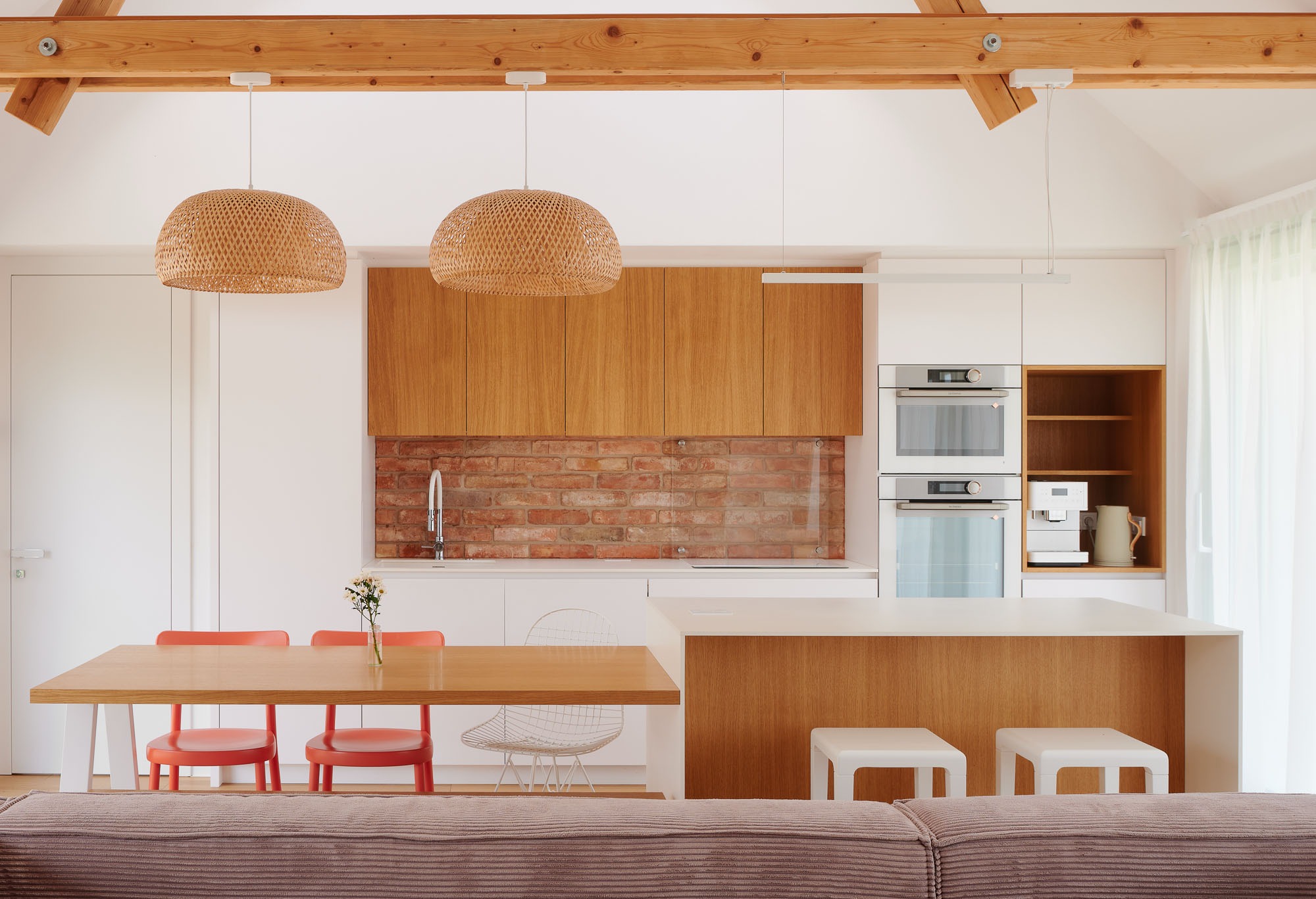 Jasna kuchnia z drewnianymi akcentami: szafki, blat, stół i lampy. Ceglana ściana nad blatem. Minimalistyczny design, nowoczesne oświetlenie, widoczne belki stropowe.