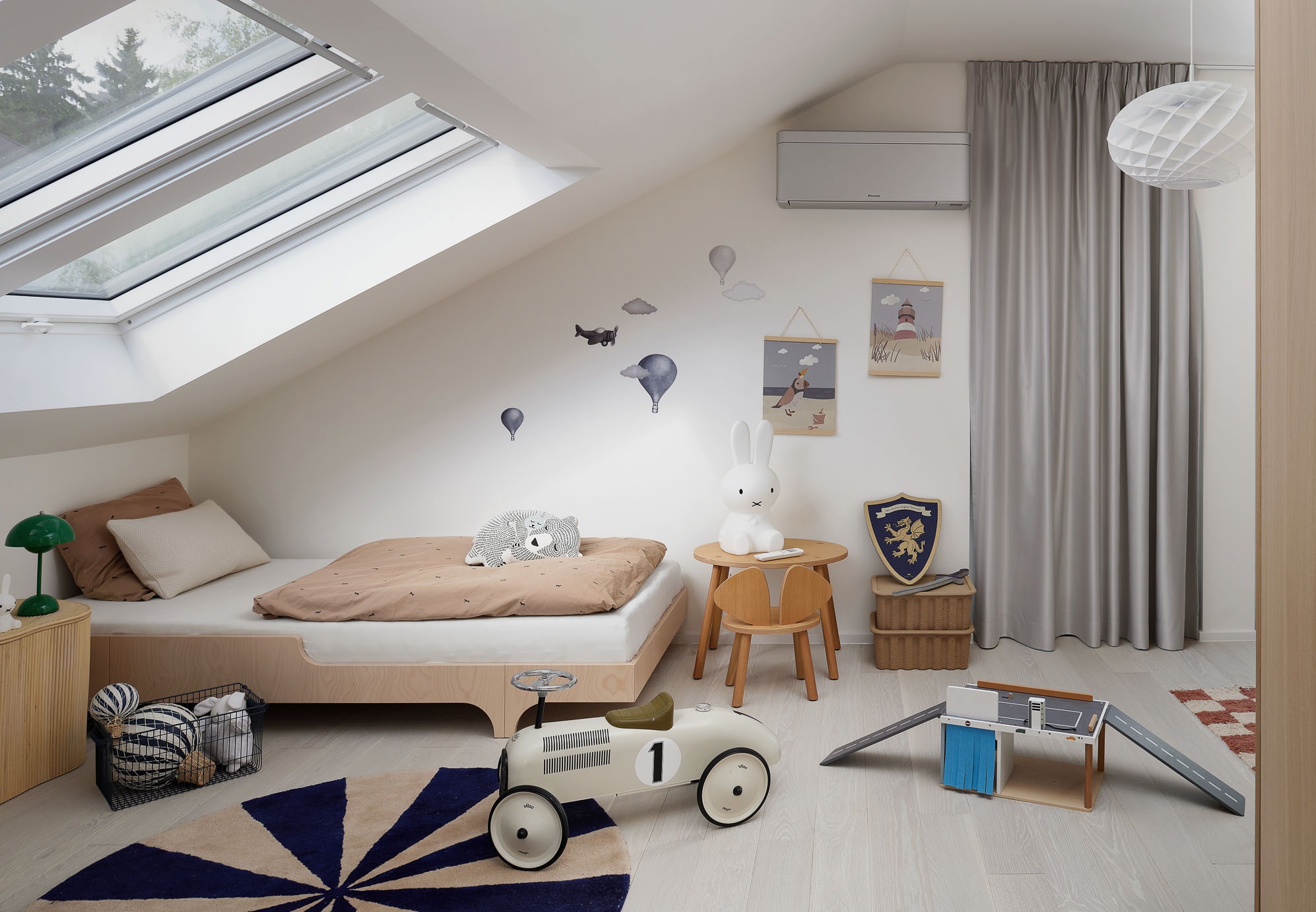Jasne poddasze z oknami dachowymi, dziecięcym łóżkiem i klimatyzatorem. Uwagę przykuwają dekoracje ścienne i zabawki, tworząc przytulny pokój.