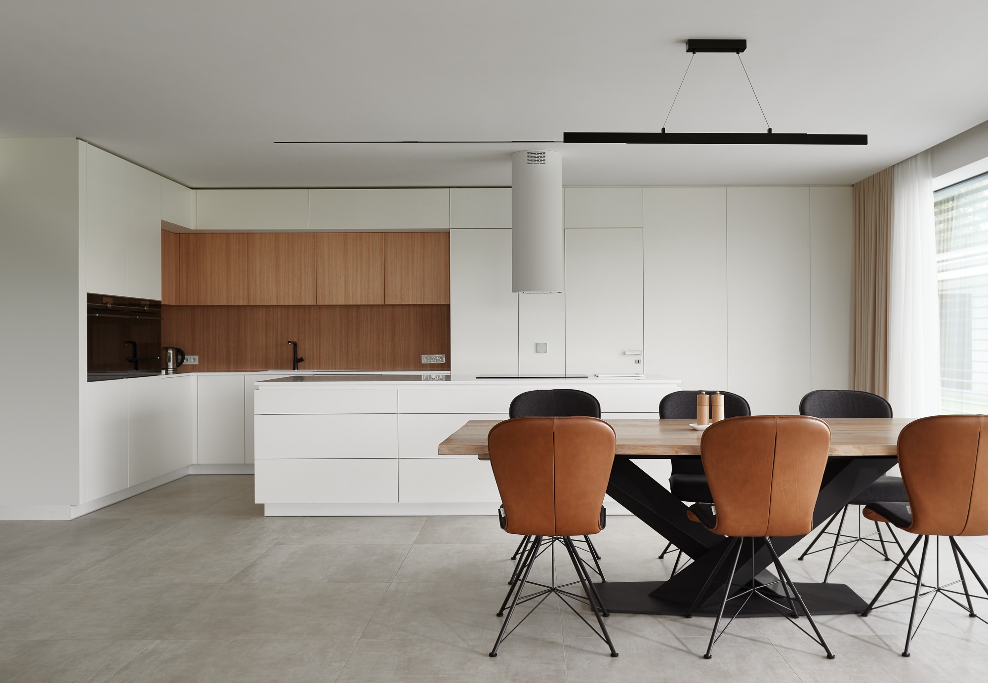 Nowoczesna kuchnia z drewnianymi akcentami i stołem jadalnym. Minimalistyczny design, jasne kolory, geometryczne lampy. Widok na okno z zasłonami.